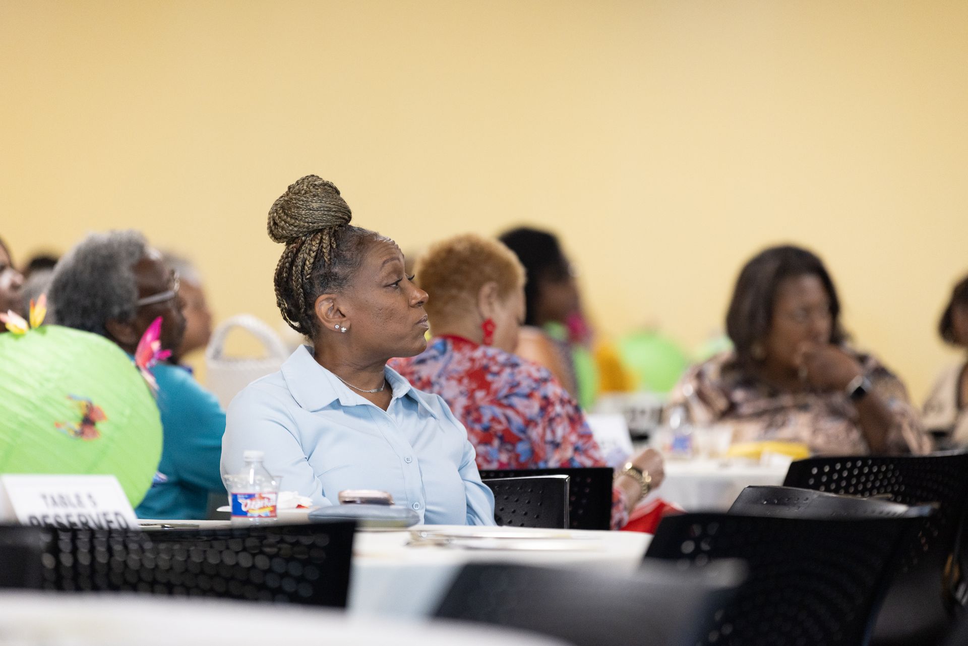 Attendees at a conference listening, seated at round tables. A woman in a light blue shirt is the focus.