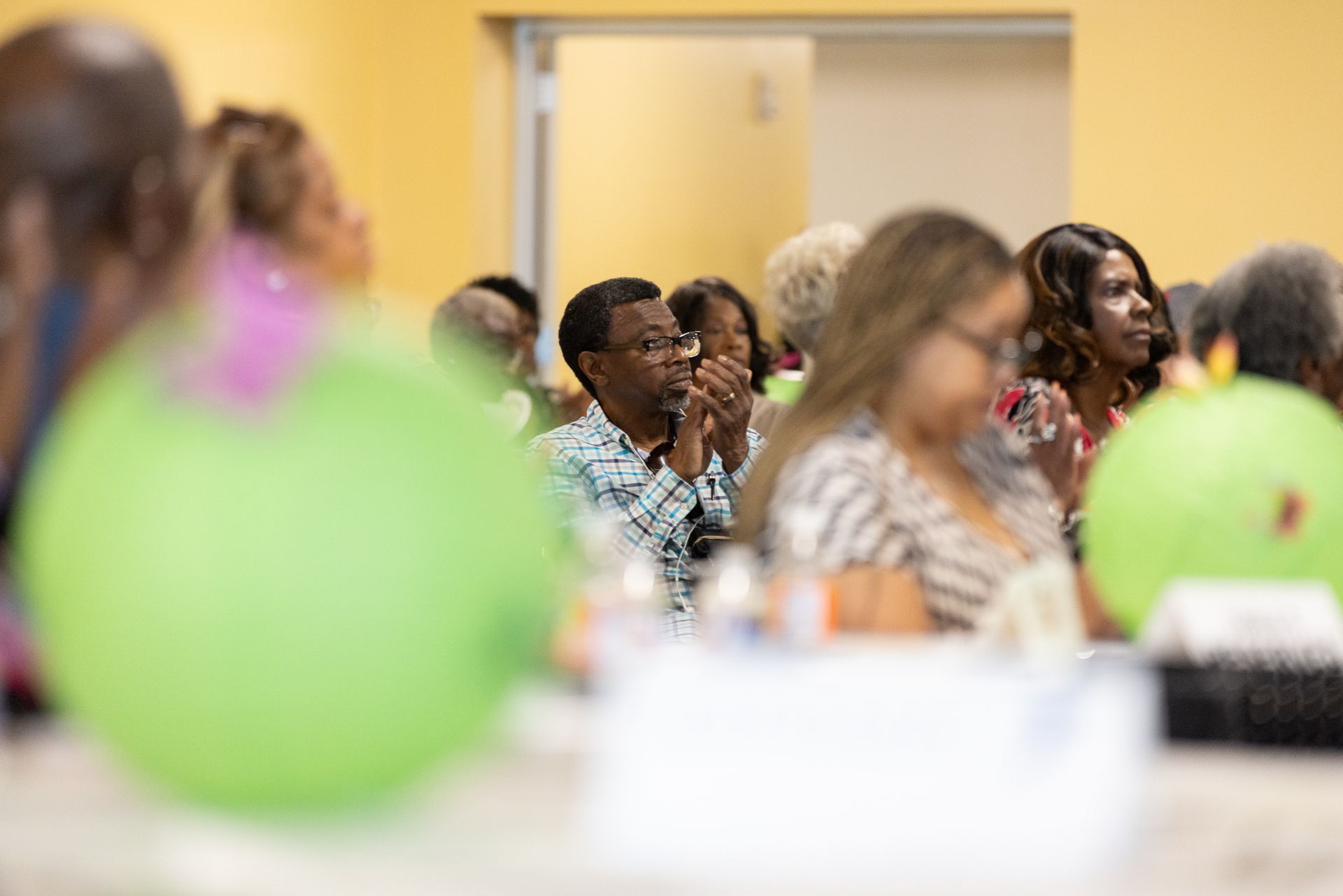 People listening attentively at an event, with green balloons in the foreground.