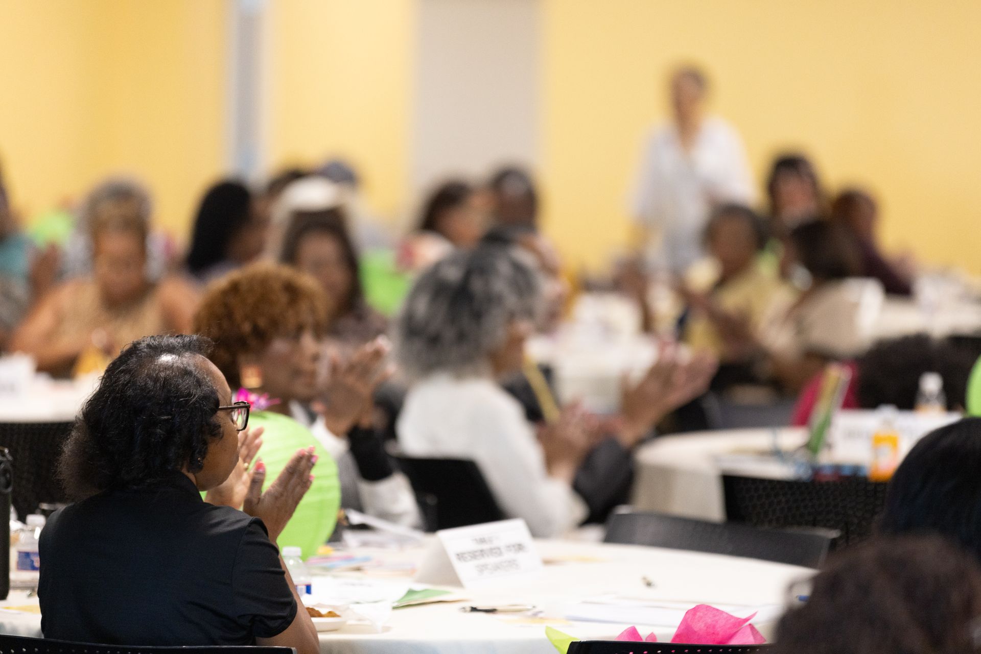 People at tables clapping in a conference room. Yellow walls, blurred background, some smiling.