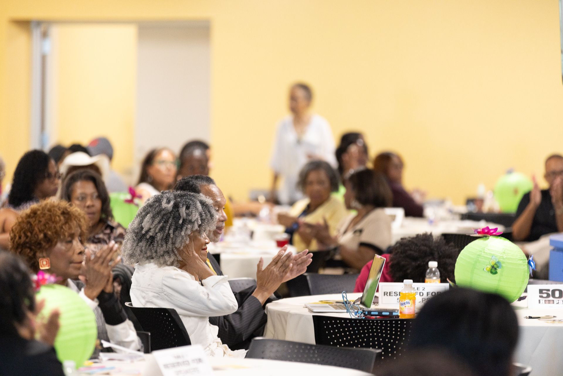 People clapping at tables in a room with a woman in the background. Green balloons on tables.