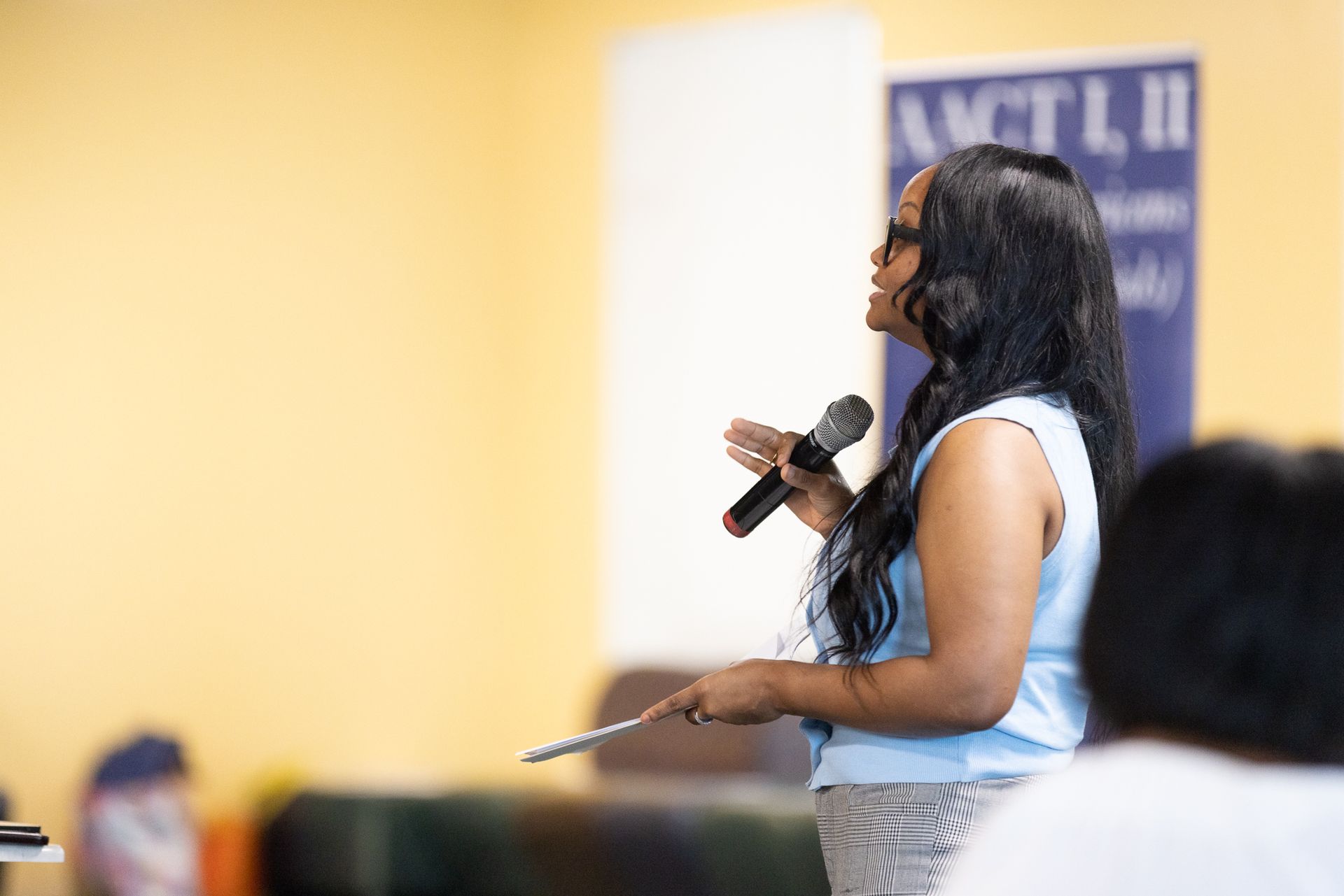 Woman speaking into a microphone, gesturing with a pen. Light blue top, standing at a podium in a yellow room.