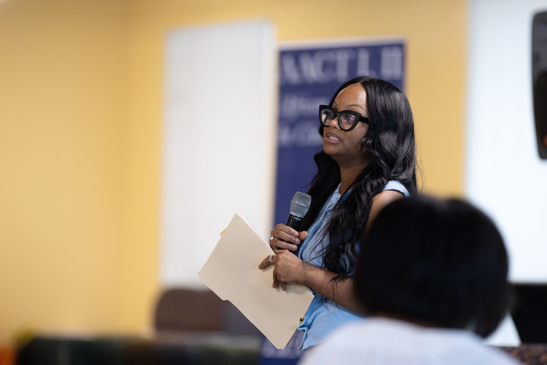 Woman with glasses speaking into a microphone, holding a folder, in a room with a banner behind her.