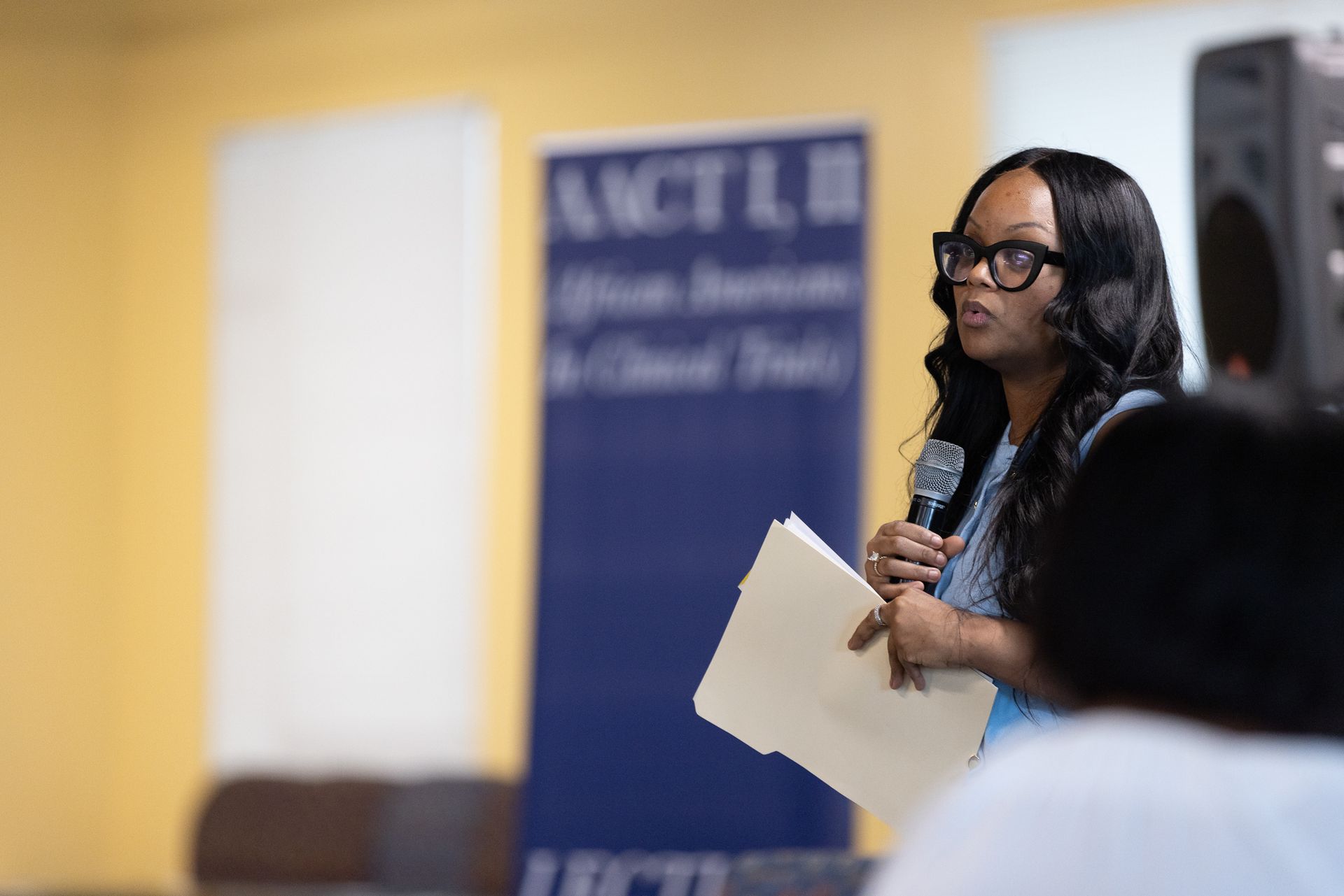 Woman speaking into microphone, holding files, in front of a banner.