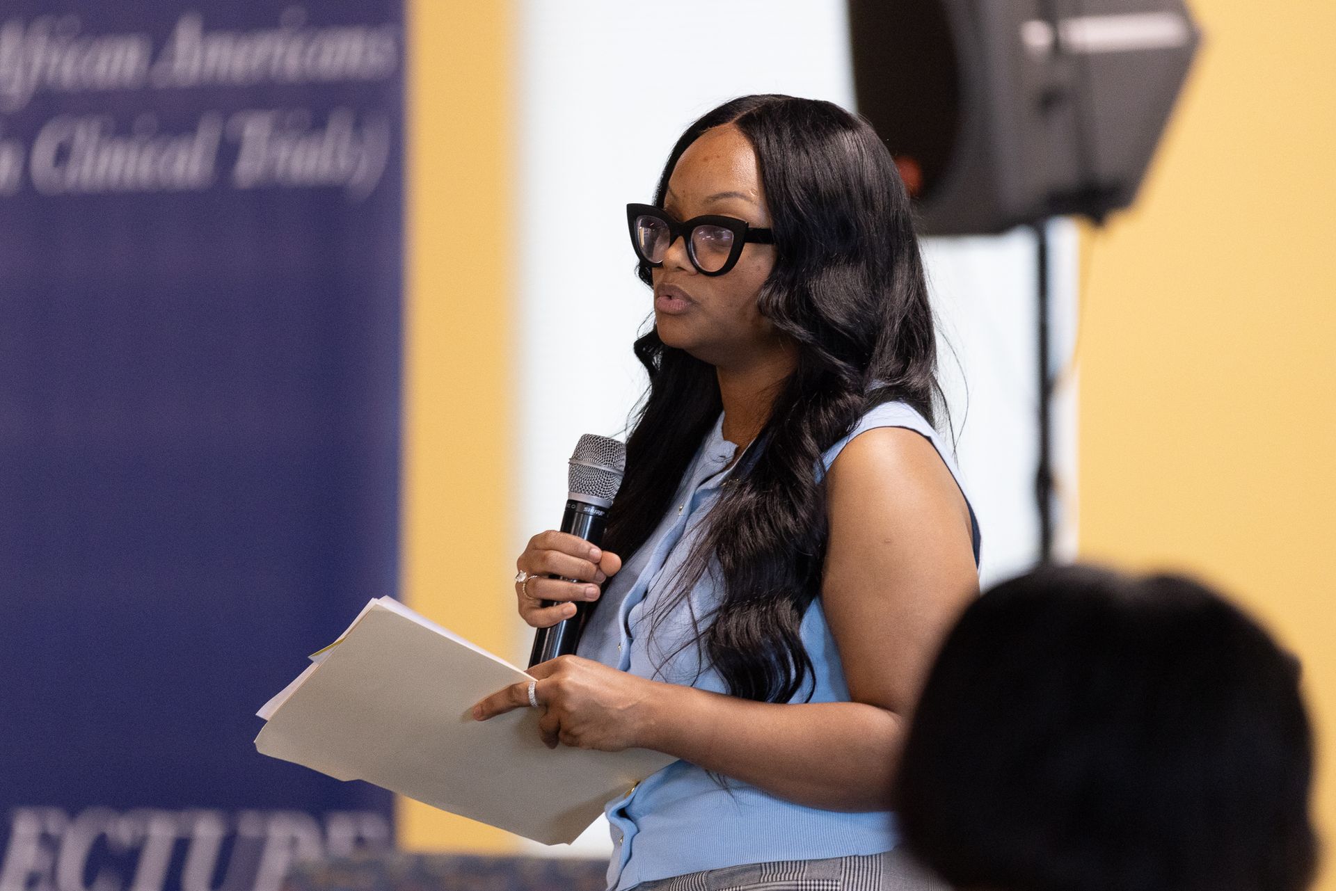 Woman speaking into a microphone, holding papers. Blue shirt, large glasses, in front of a blue banner.