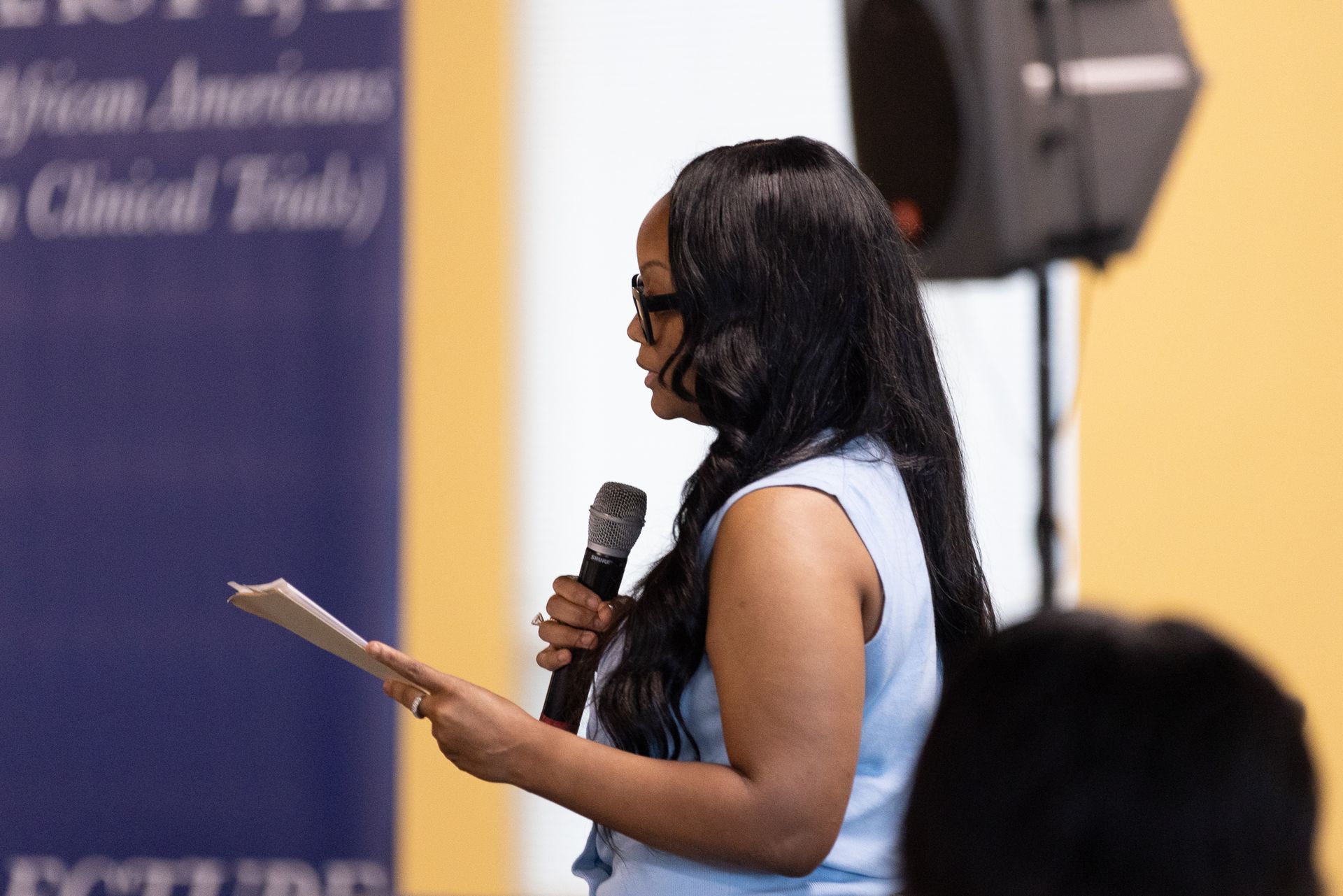 Woman speaking at lectern, holding paper and microphone, in front of a blue banner and yellow wall.