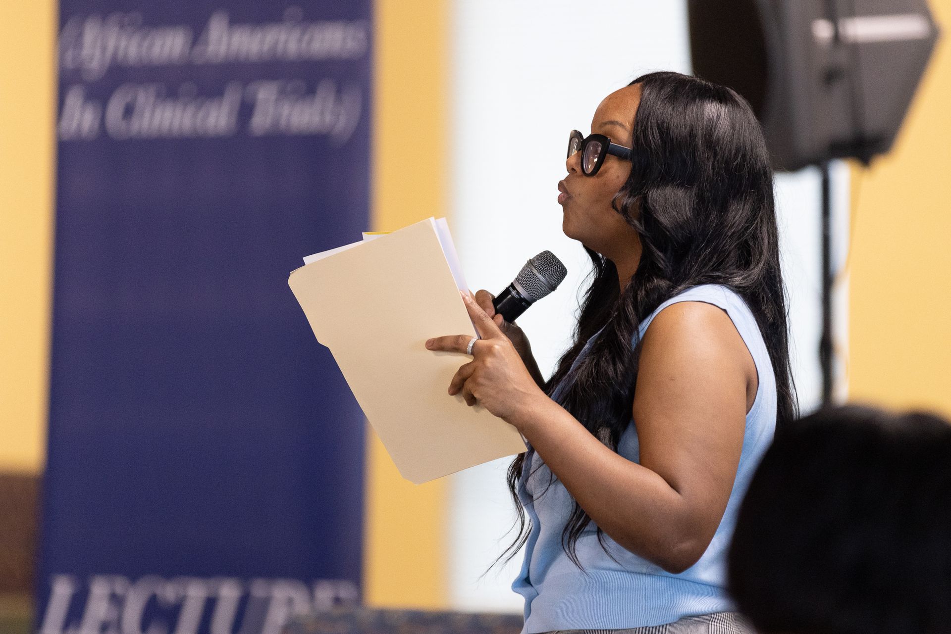Woman speaking into a microphone, holding papers, at a lecture, with a banner behind her.