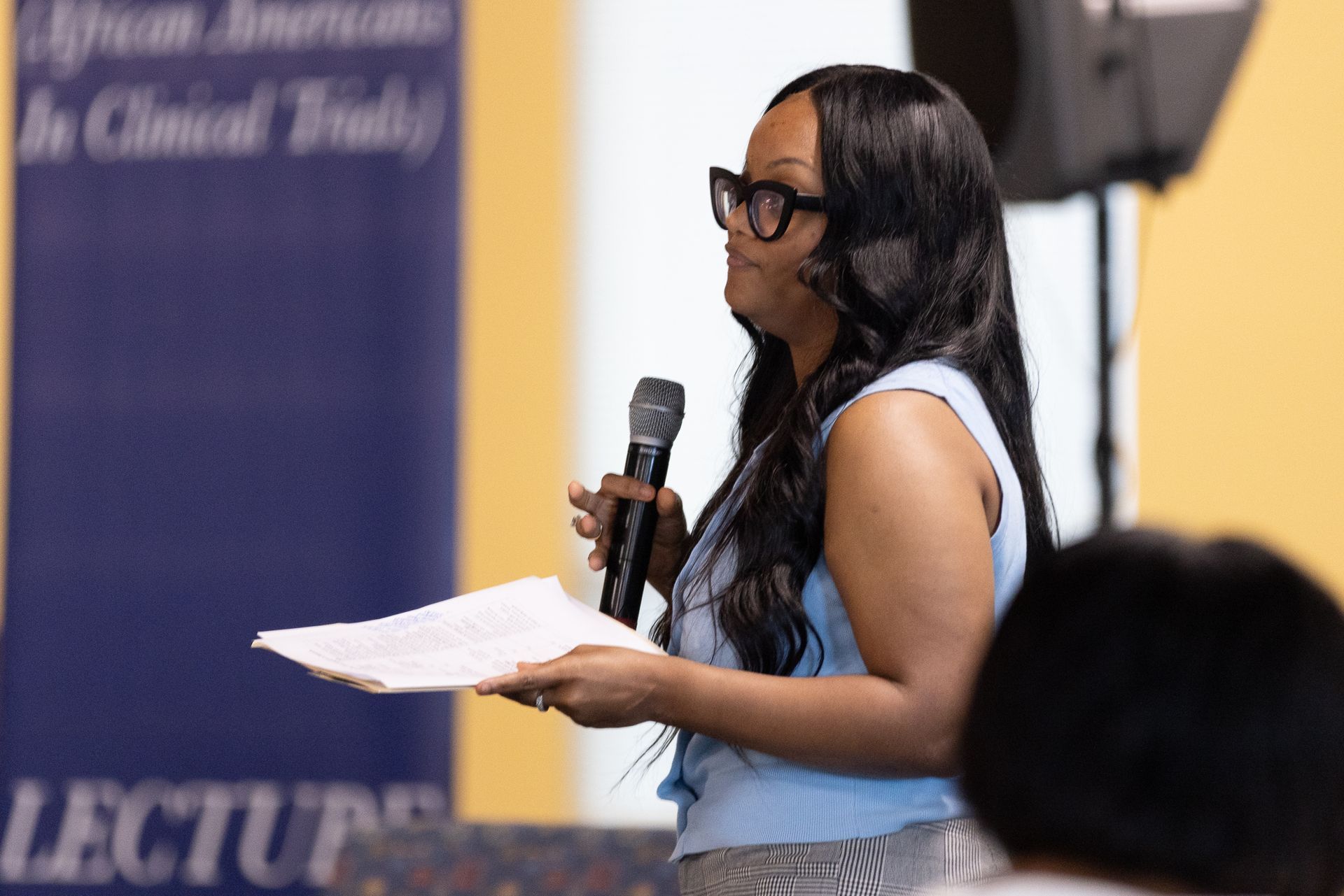 Woman speaking into microphone, holding papers, in front of a banner about African Americans in clinical trials.