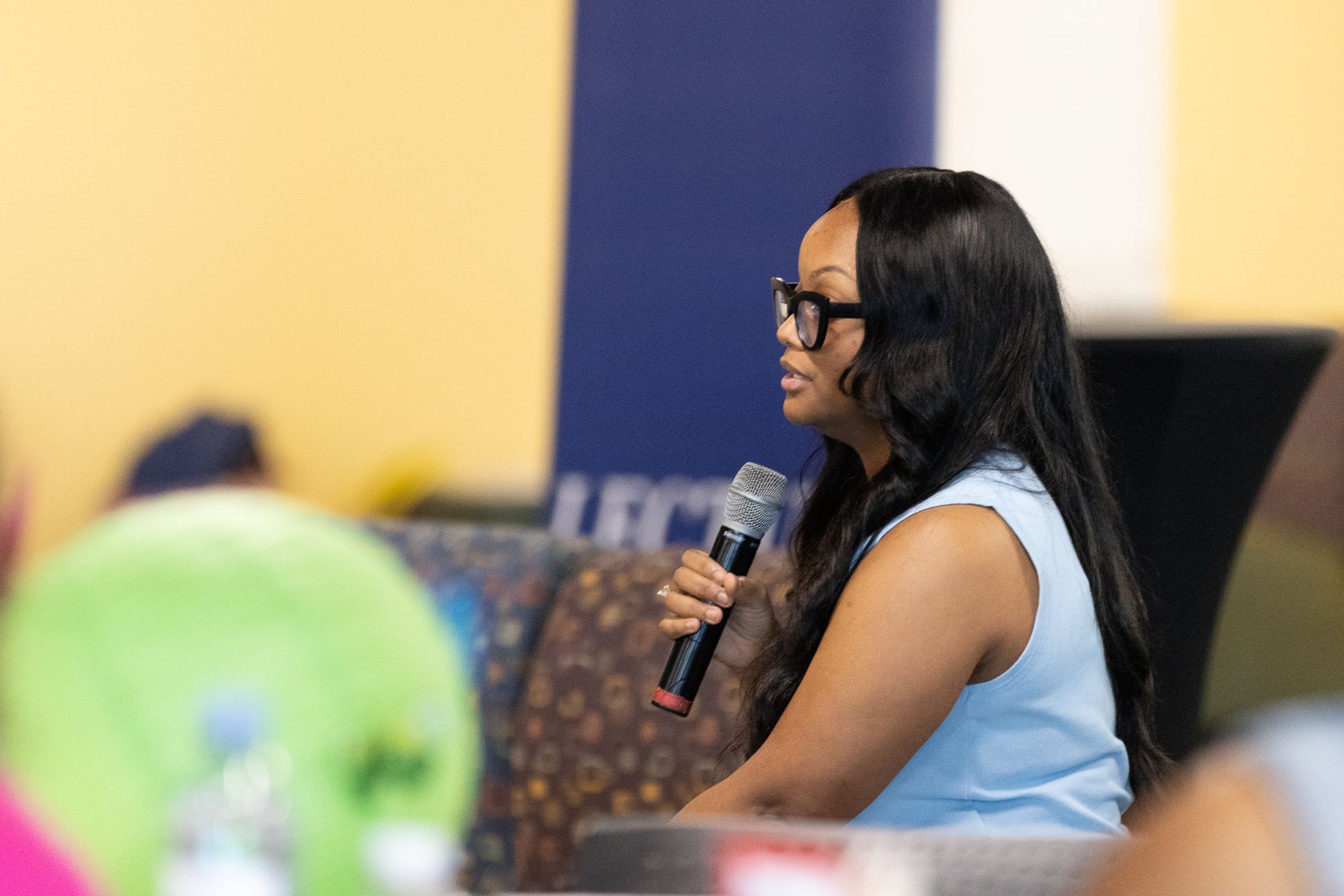 Woman speaking into a microphone, wearing glasses, blue top. Seated at an event.