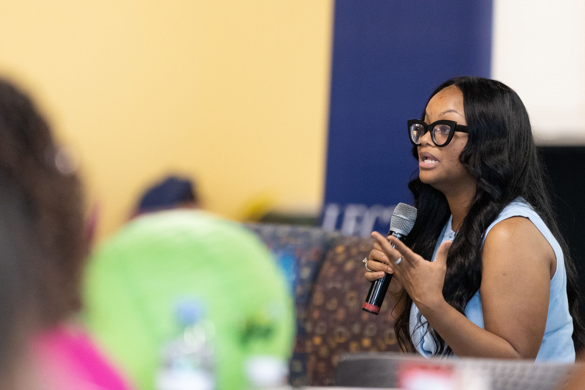 Woman speaking into microphone, wearing glasses, in a brightly lit room.