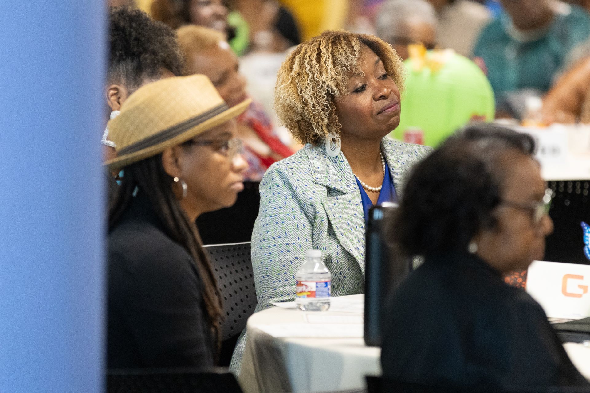 Two women at a table at an event, one wearing a hat, the other in a blazer, listening.