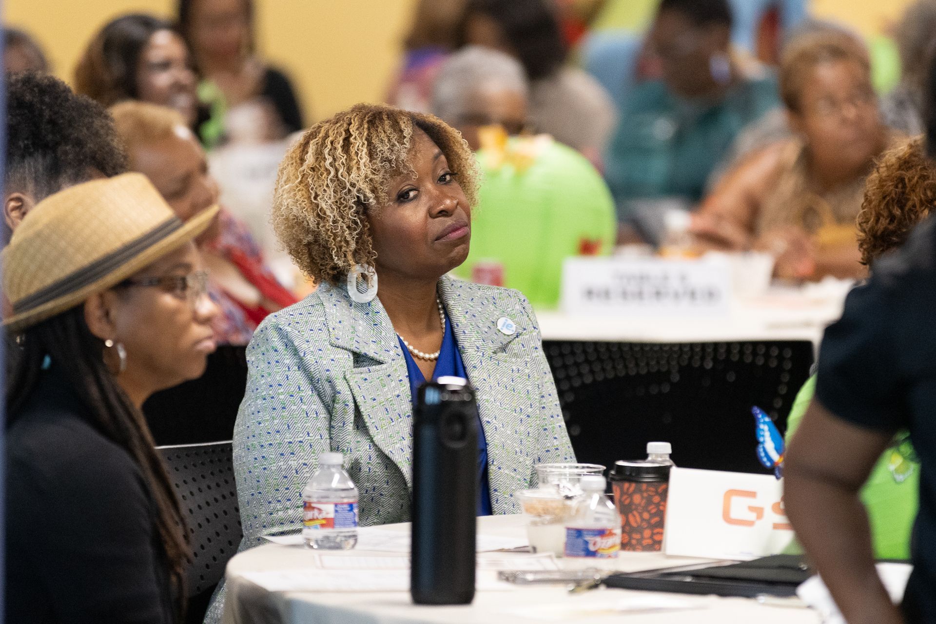 Woman in a light blue blazer at a conference, looking to the side; people in the background.