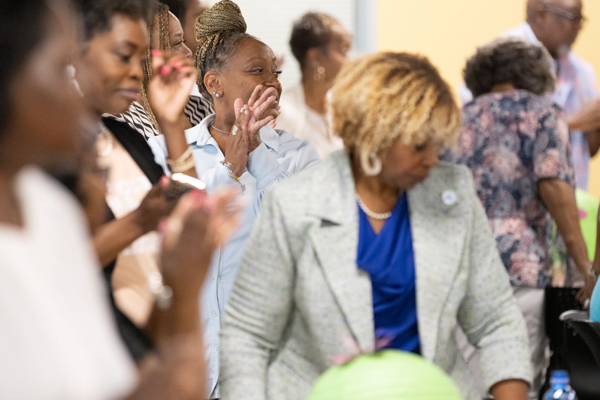 People in a room, some clapping. Woman in a gray blazer and blue top walks toward foreground.