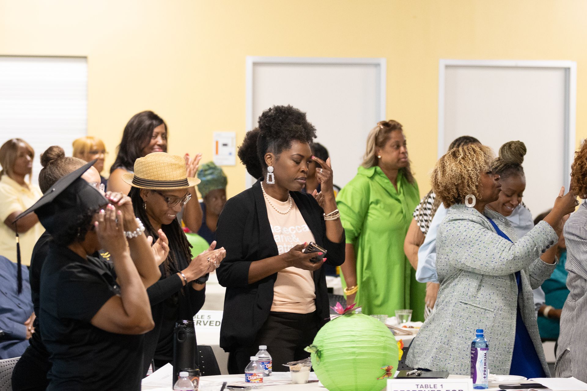 Group of people inside a room, some clapping, some looking down. Yellow and white walls, tables set with items.