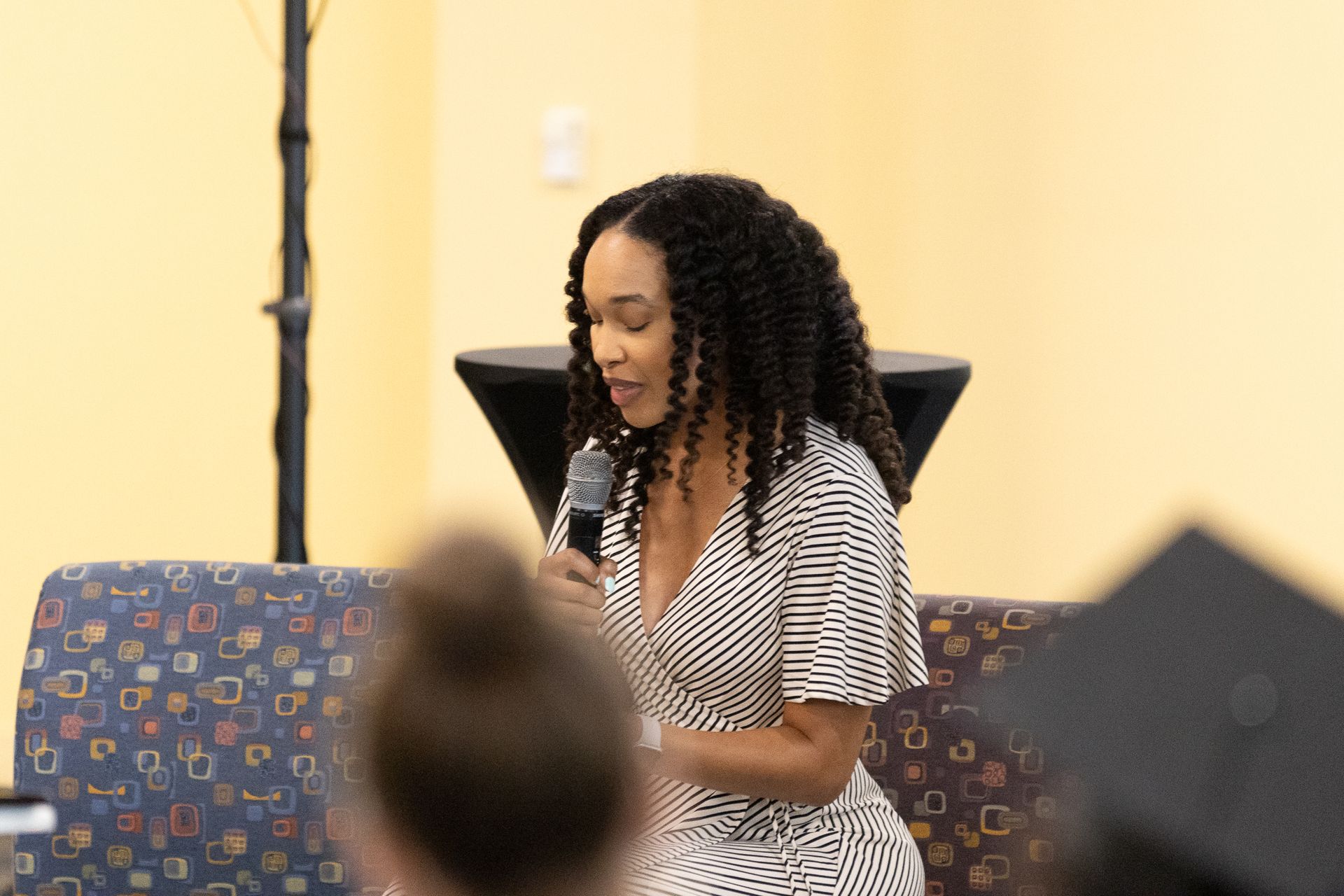 Woman speaking into a microphone, sitting on a patterned couch. A table and person's back are in the foreground.