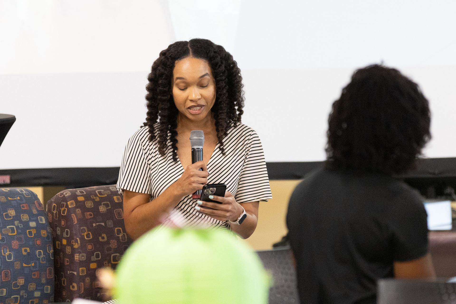 Woman speaking into a microphone, holding a phone, in front of a projection screen.
