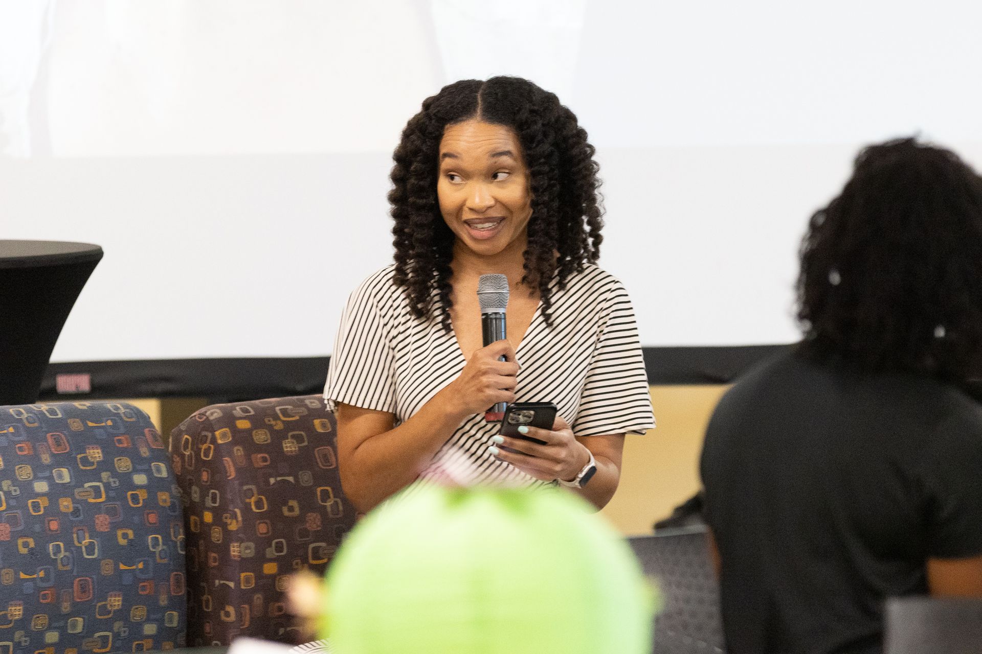 Woman speaking into a microphone, holding a phone, in a room with a screen and audience.