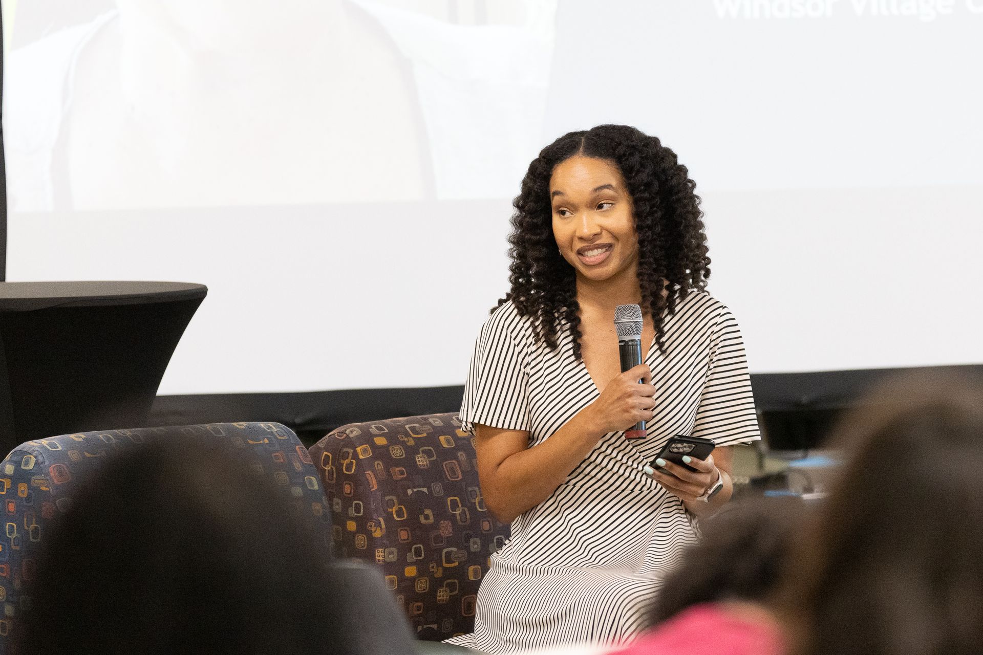 Woman speaking into a microphone, holding a phone, sitting at a table in front of a screen.