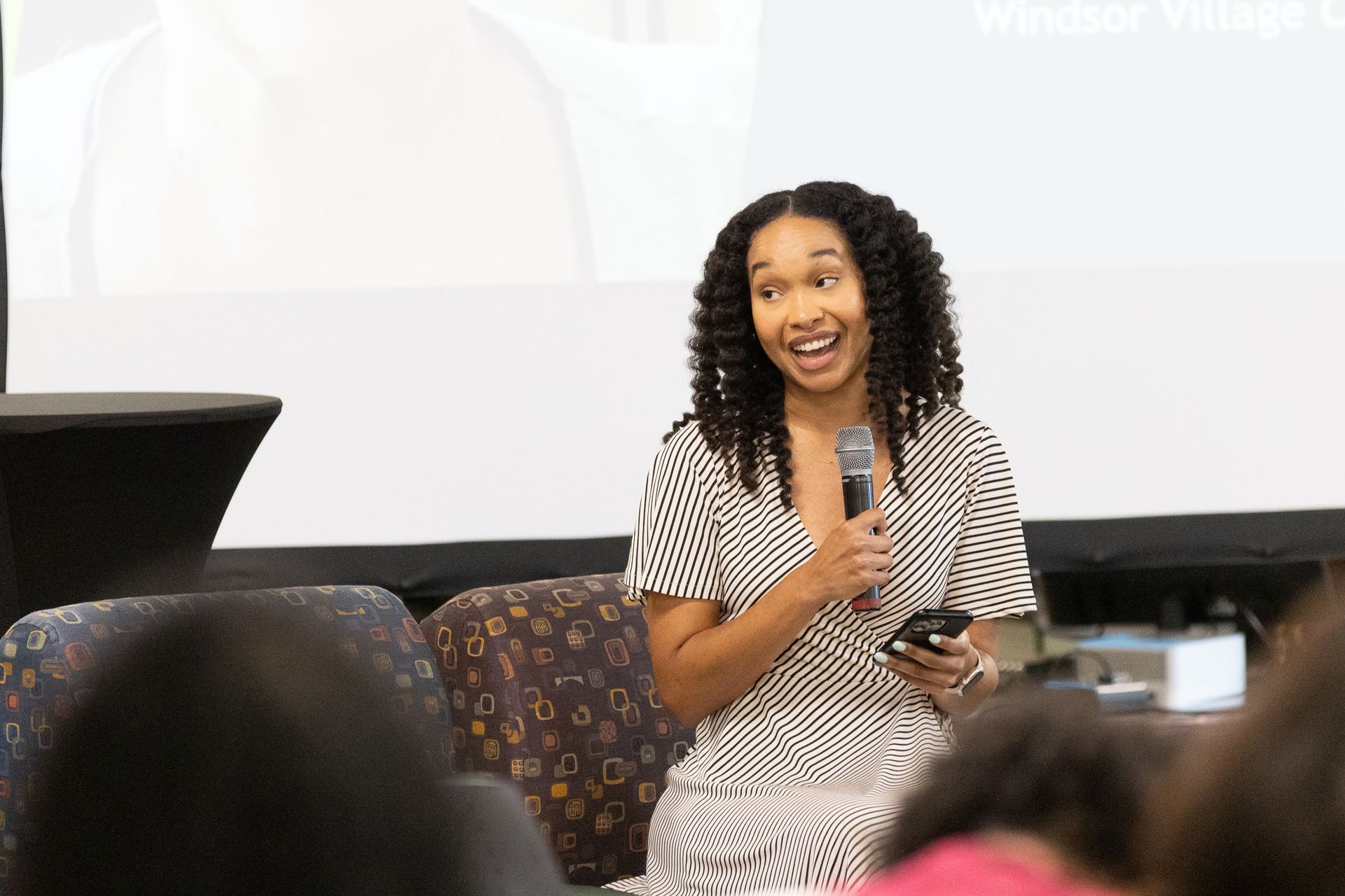 Woman with curly hair speaking into a microphone, holding a phone, sitting on stage, smiling.