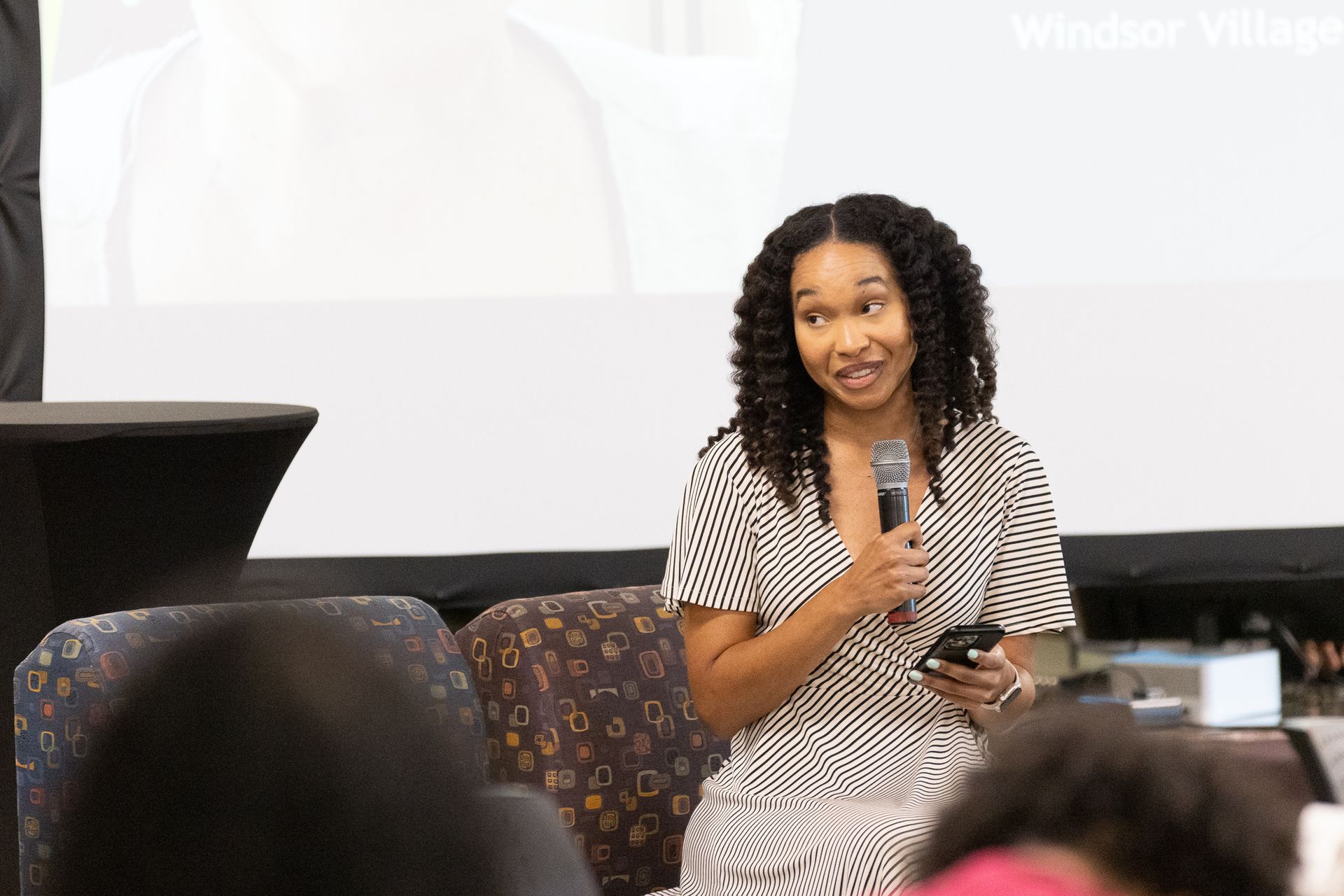 Woman with microphone, smiling, in front of a screen, holding a small device, on a stage.