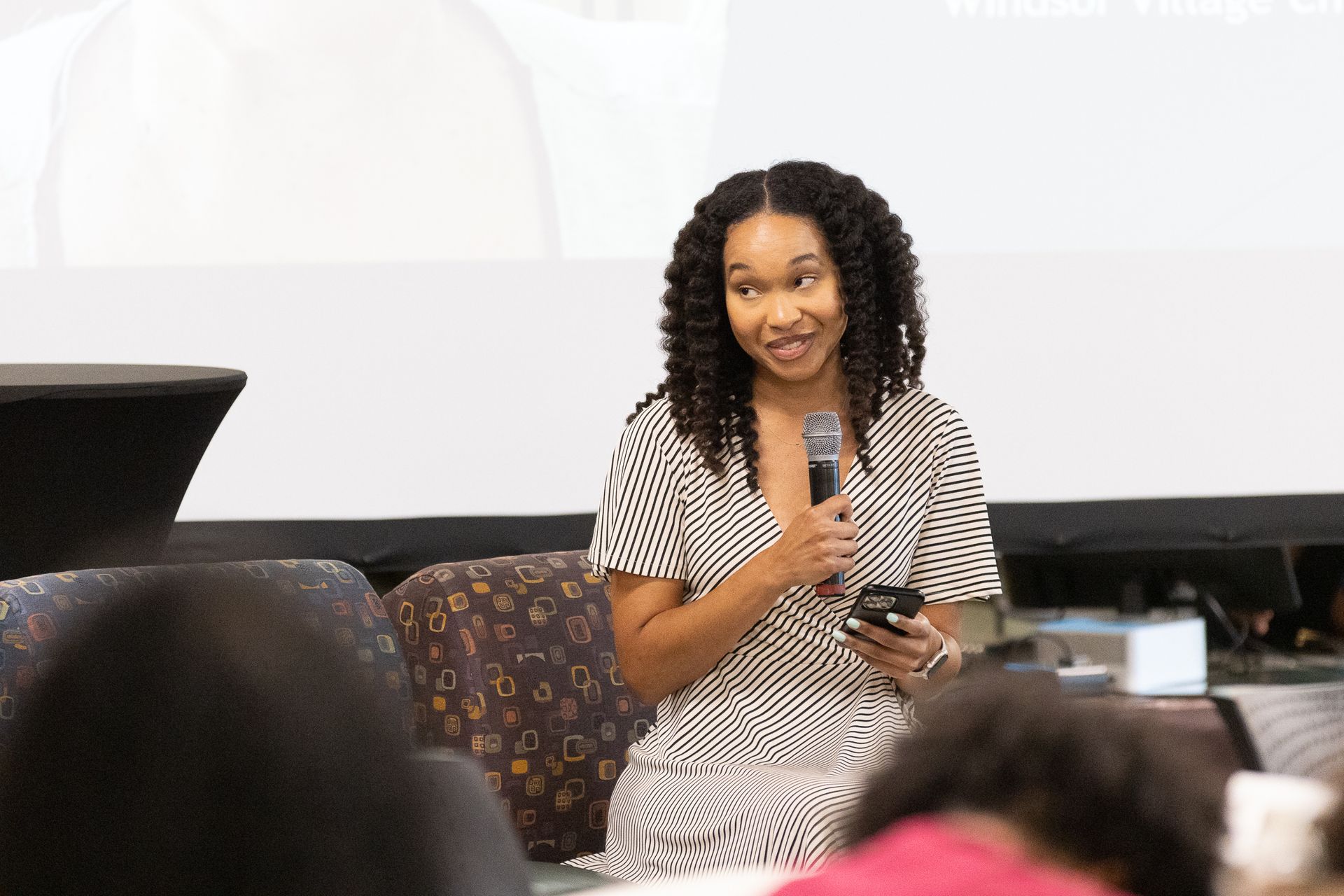 Woman speaking into a microphone, holding a phone, sitting on a stage in front of an audience.