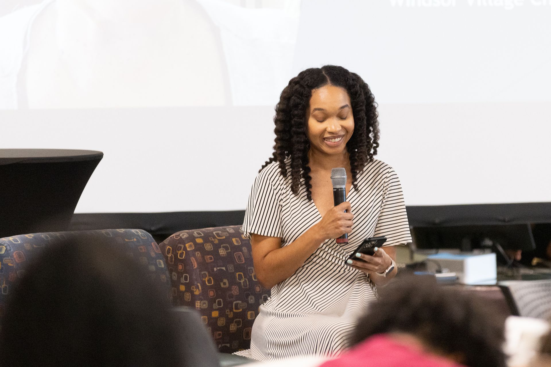Woman speaking into a microphone, holding a phone, sitting on stage in front of a screen.