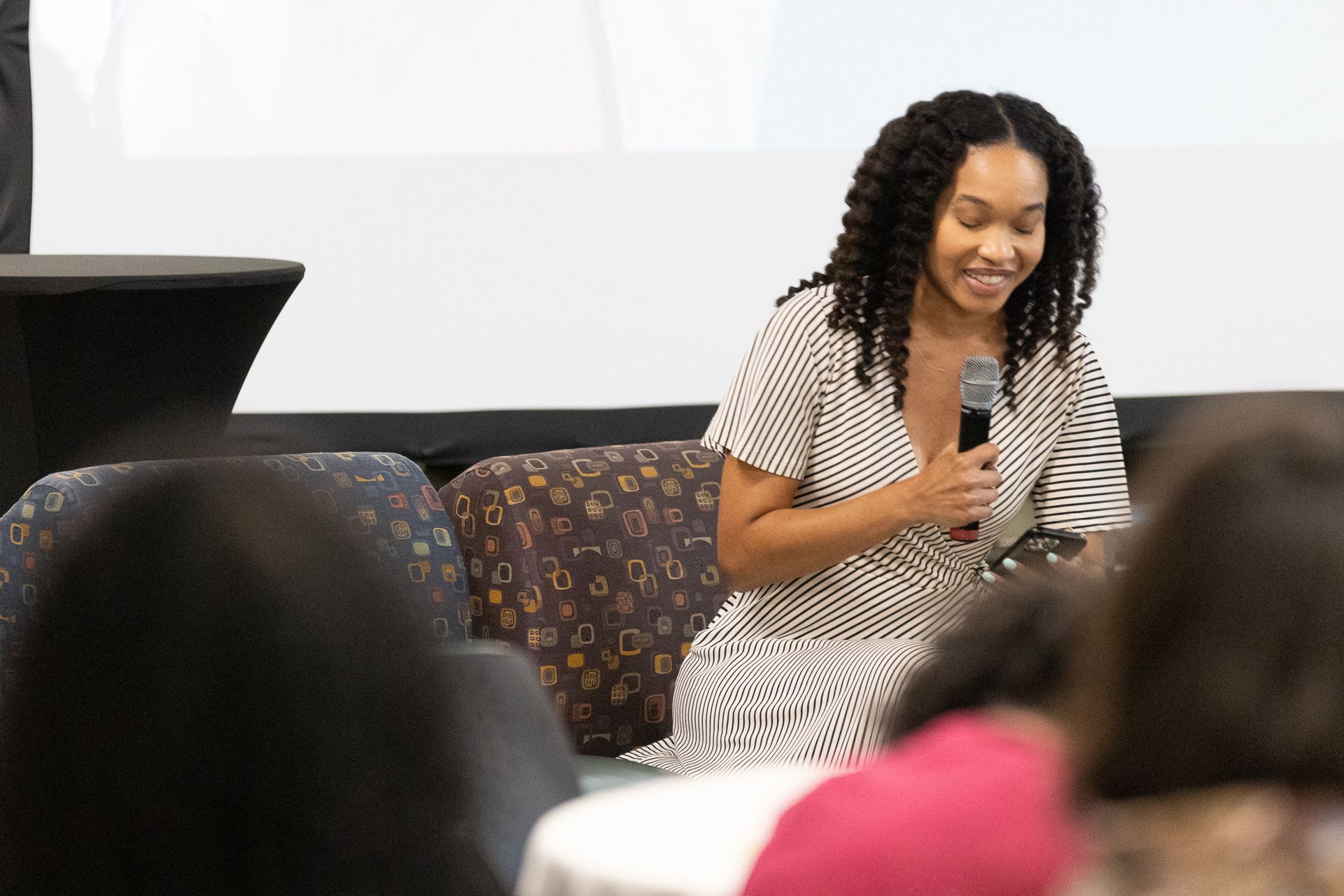Woman with microphone speaks while seated on a couch in front of an audience.