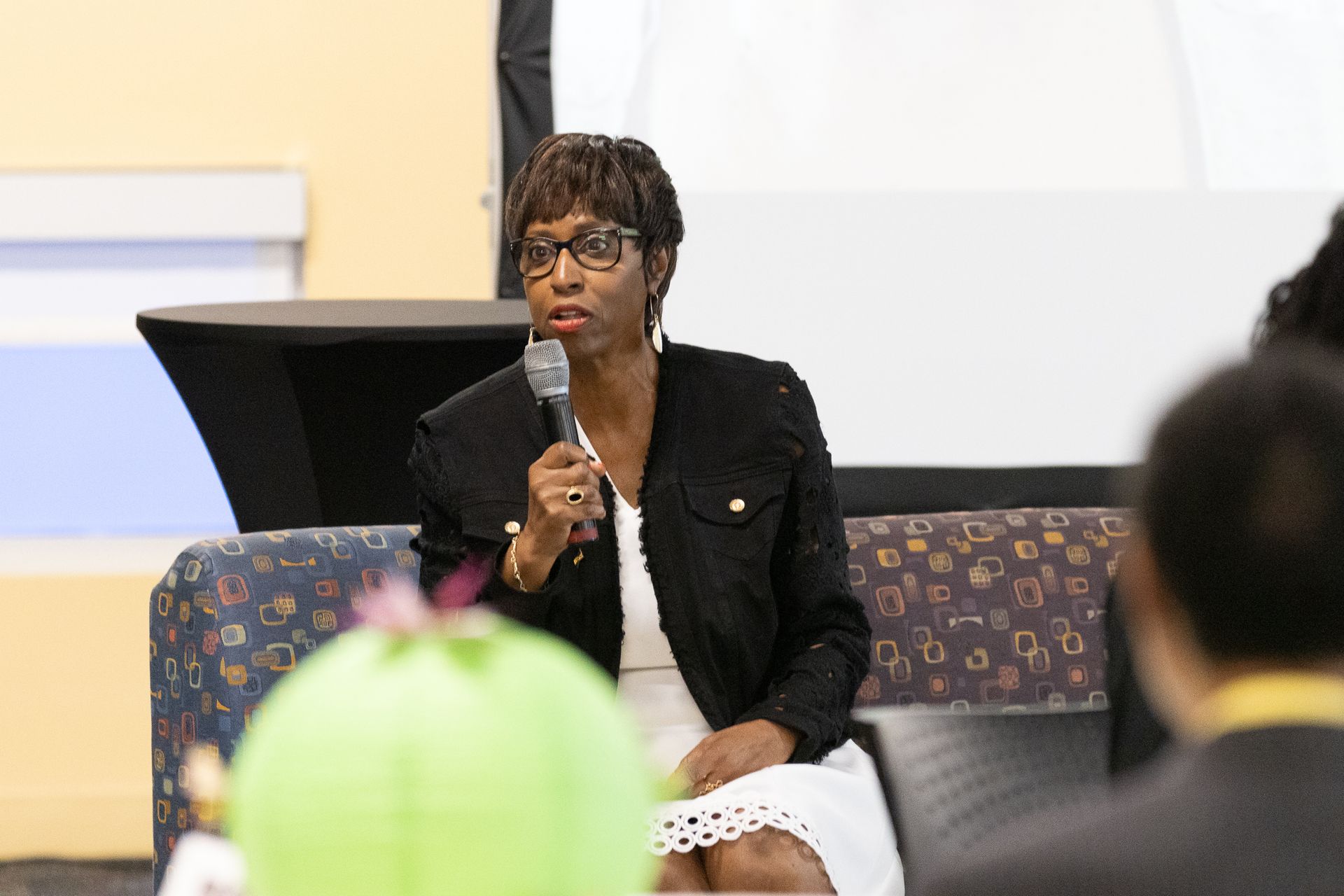 Woman speaking into a microphone, seated on a patterned sofa in a conference setting.