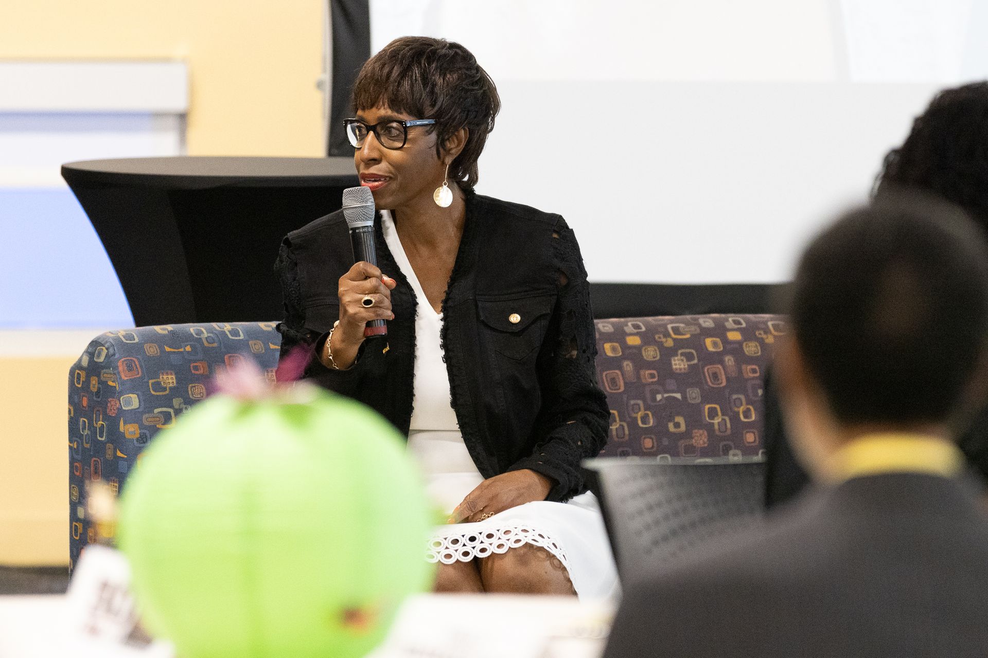Woman speaking into a microphone, sitting on a stage. She wears a black jacket and white dress.