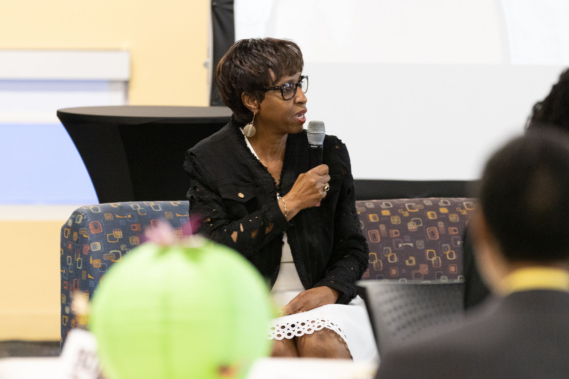 Woman in black jacket speaks into microphone, seated on patterned couch. A green paper lantern is in the foreground.