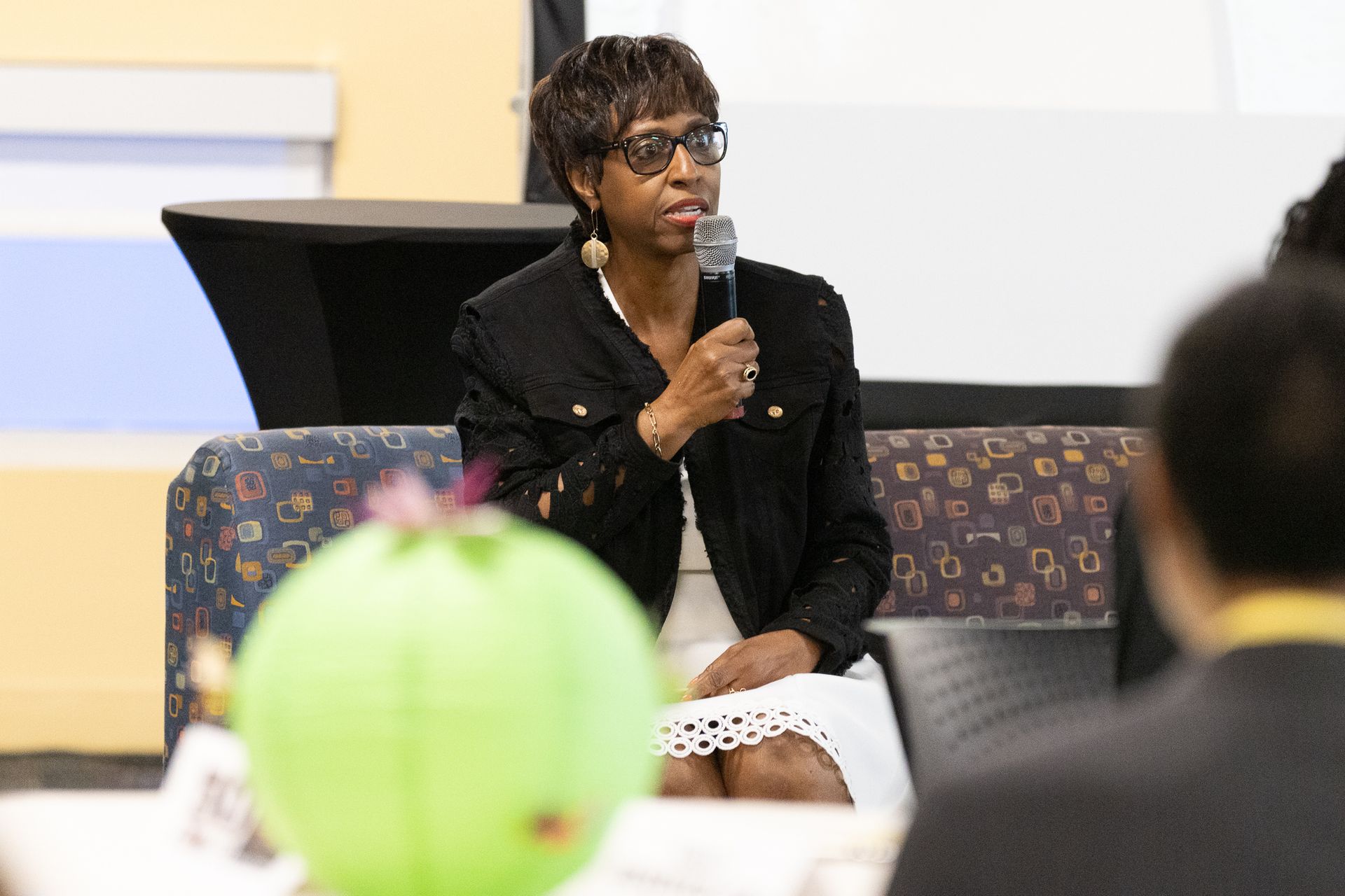 Woman speaking into microphone at an event, wearing glasses and a black jacket, green centerpiece in foreground.