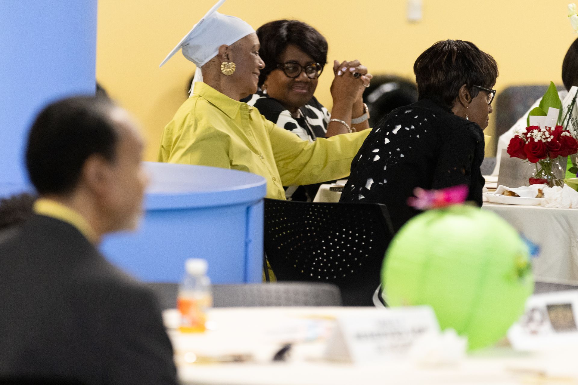 A woman in a yellow shirt and graduation cap raises her hand, celebrating at a seated event.