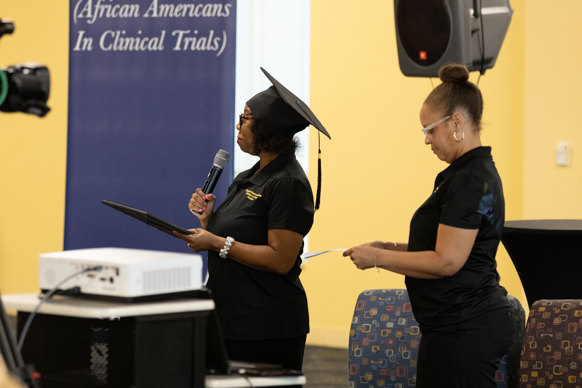 Two women at an event; one in graduation cap, holding a microphone. Blue banner visible.