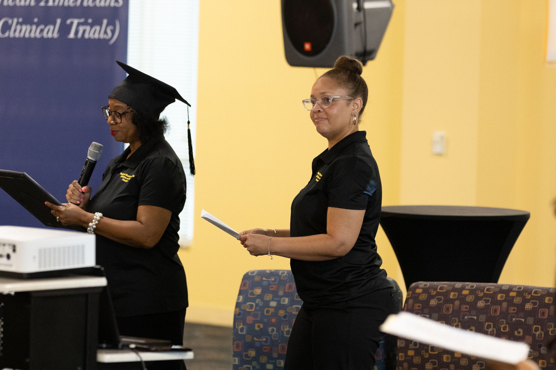 Two women in black shirts on stage, one with a graduation cap and microphone, reading. Yellow wall.