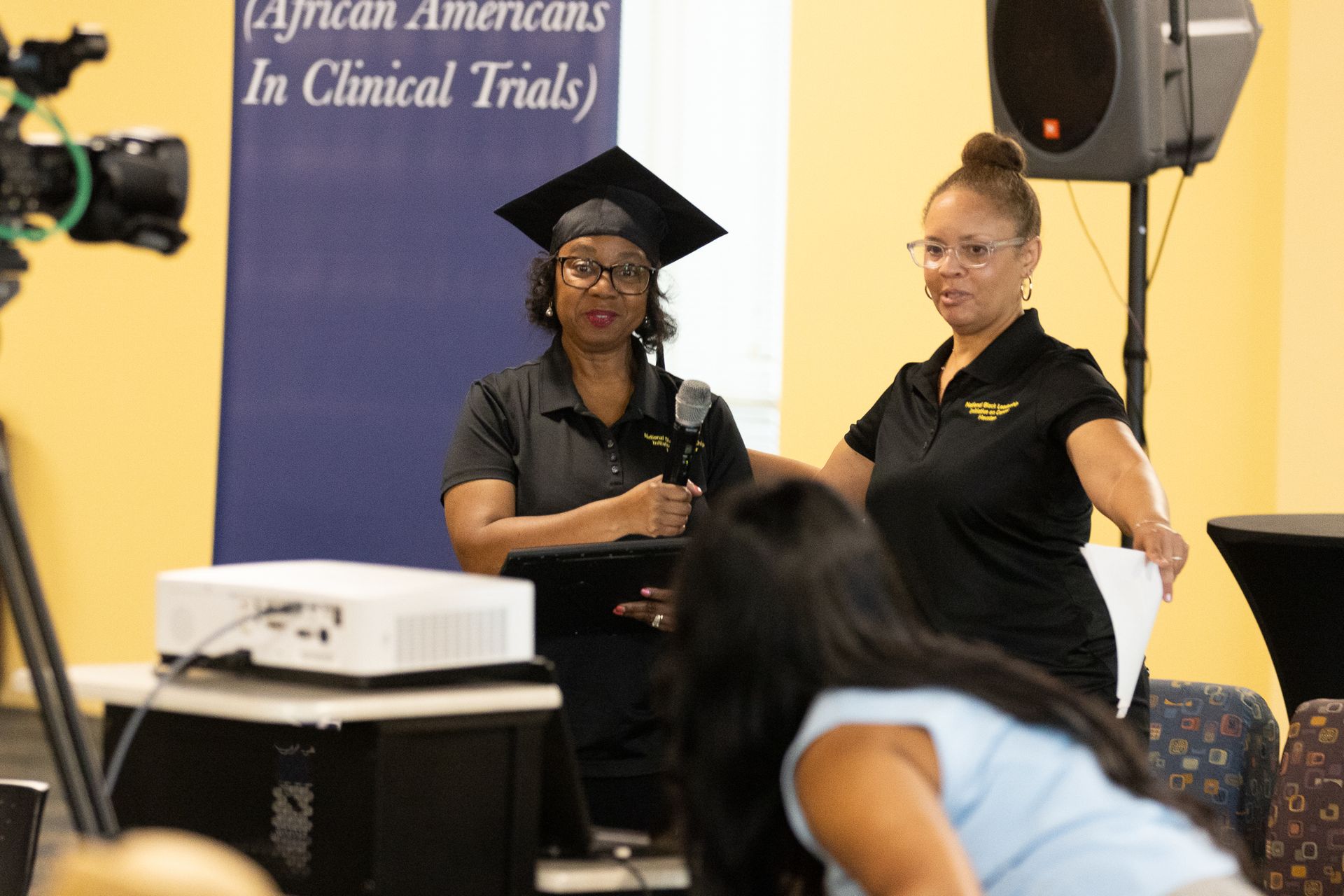 Two women at a presentation, one in a graduation cap. The banner reads 