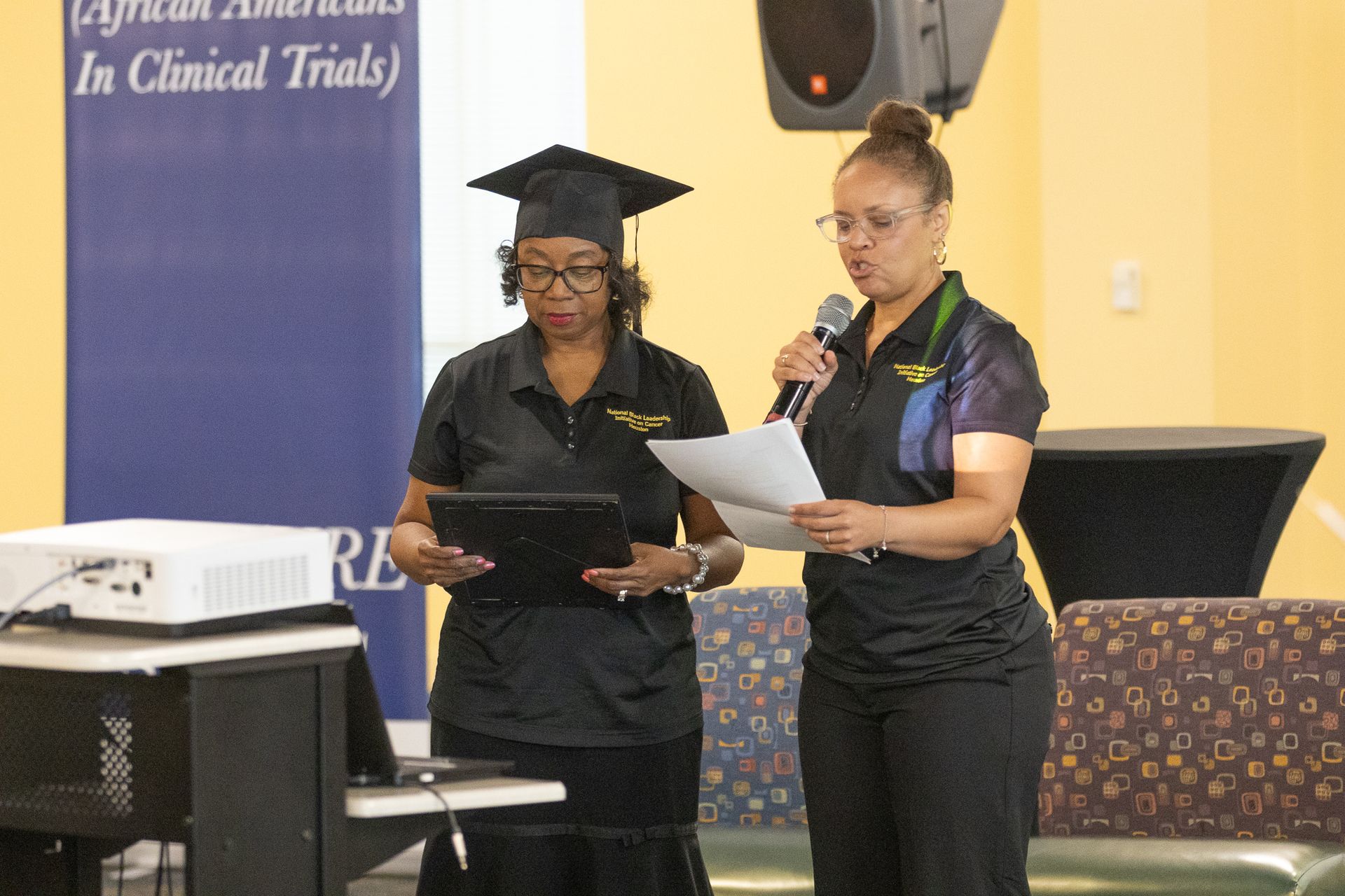 Two women presenting at a conference. One in a graduation cap, the other with a microphone.