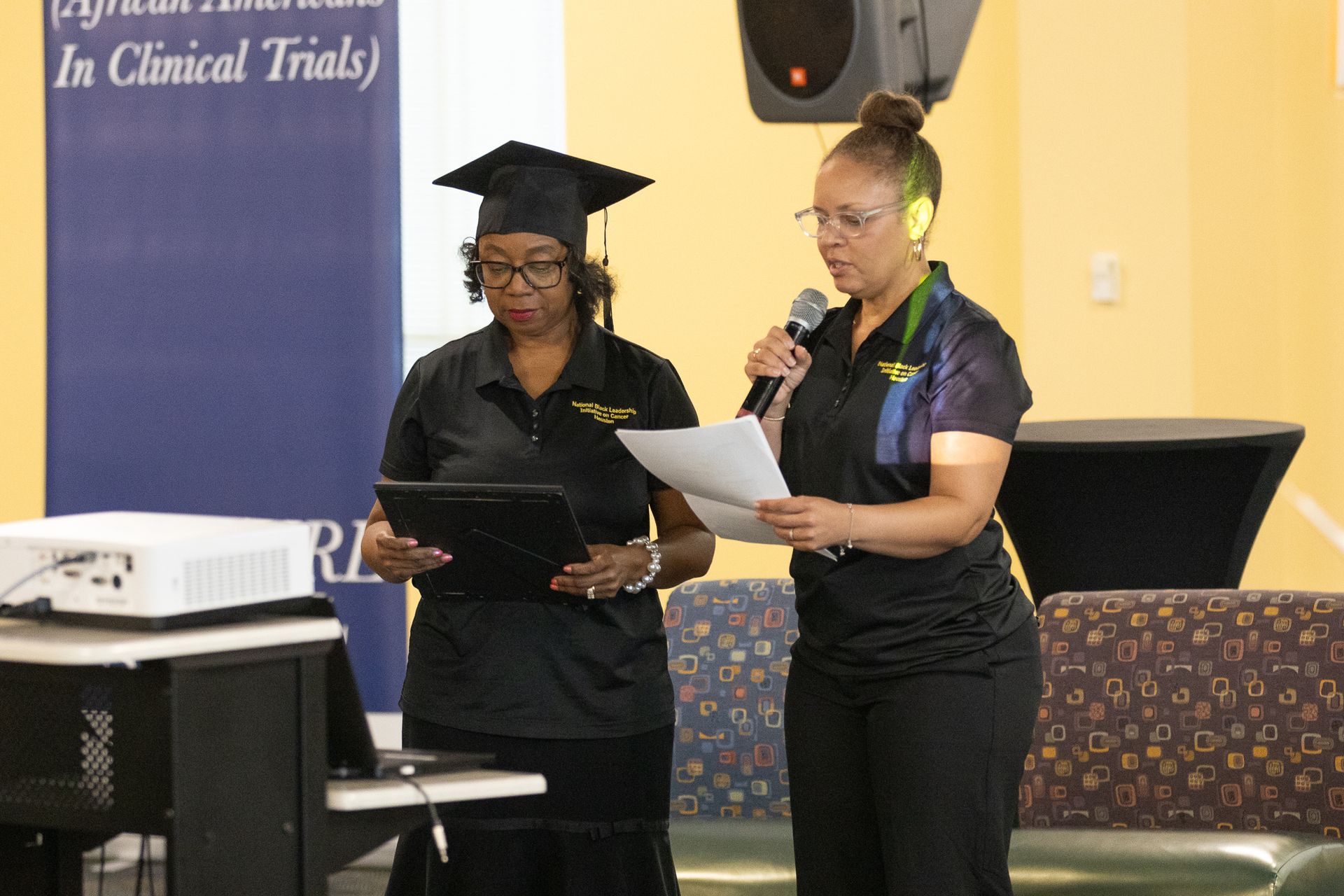 Two women at podium. One in cap and gown, other with microphone, both in black. Presentation setting.