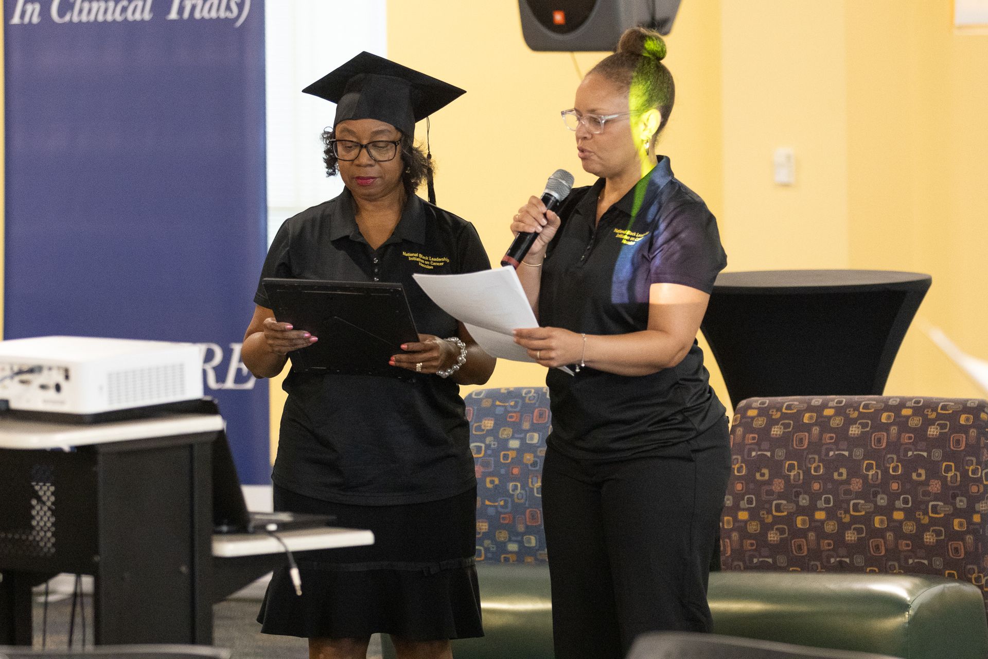 Two women at a presentation; one wears a graduation cap. They're in front of a screen and holding papers.