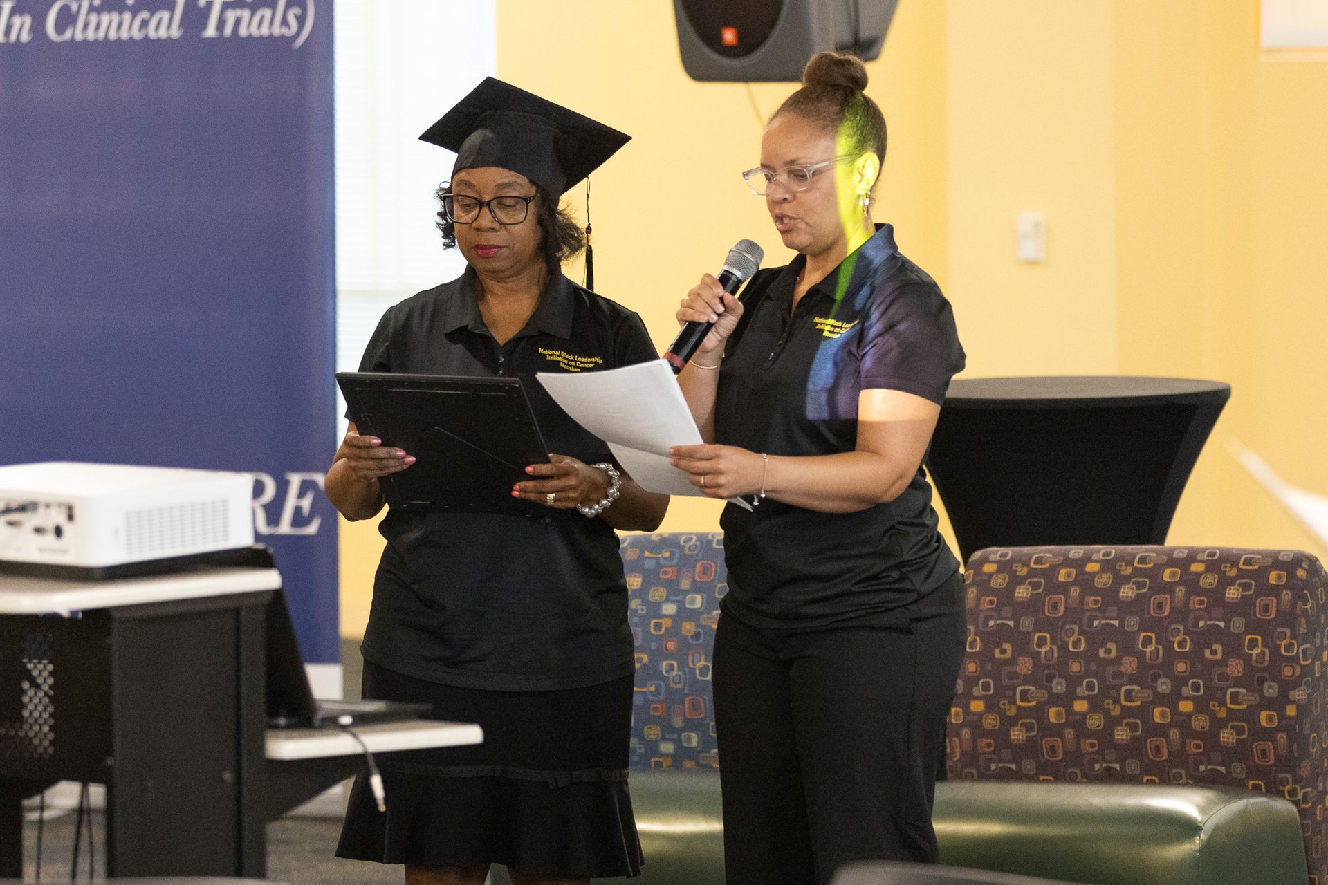 Two women at podium. One in cap and gown, other with microphone. Both reading papers.