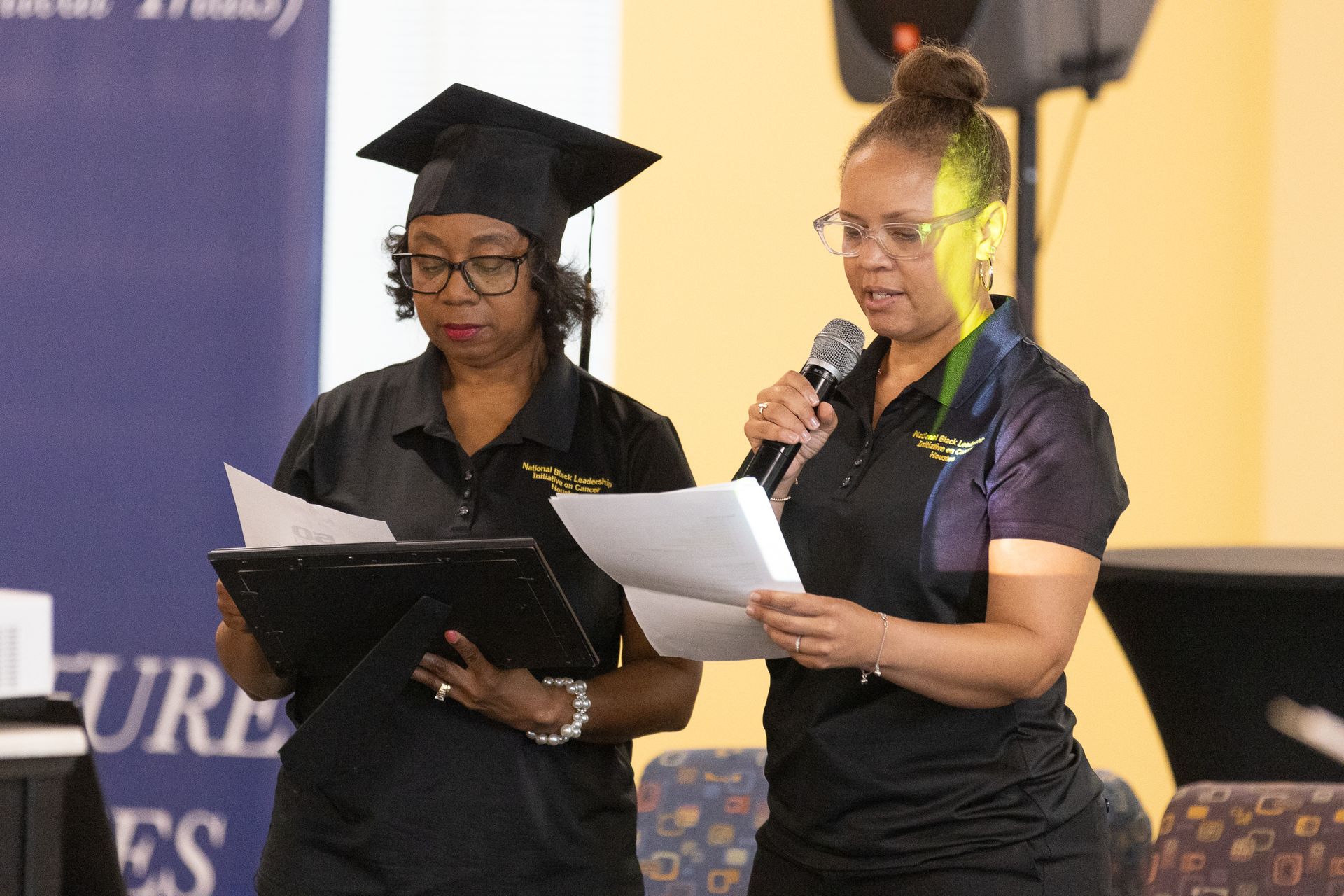 Two women at a podium, one in a cap, reading from papers. The woman on the right holds a microphone.