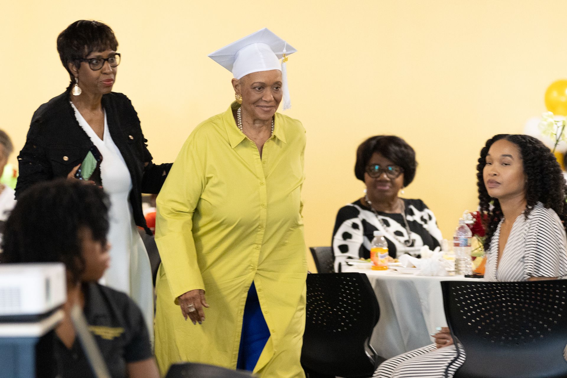 Woman in graduation cap and yellow dress walks past seated guests in a room.