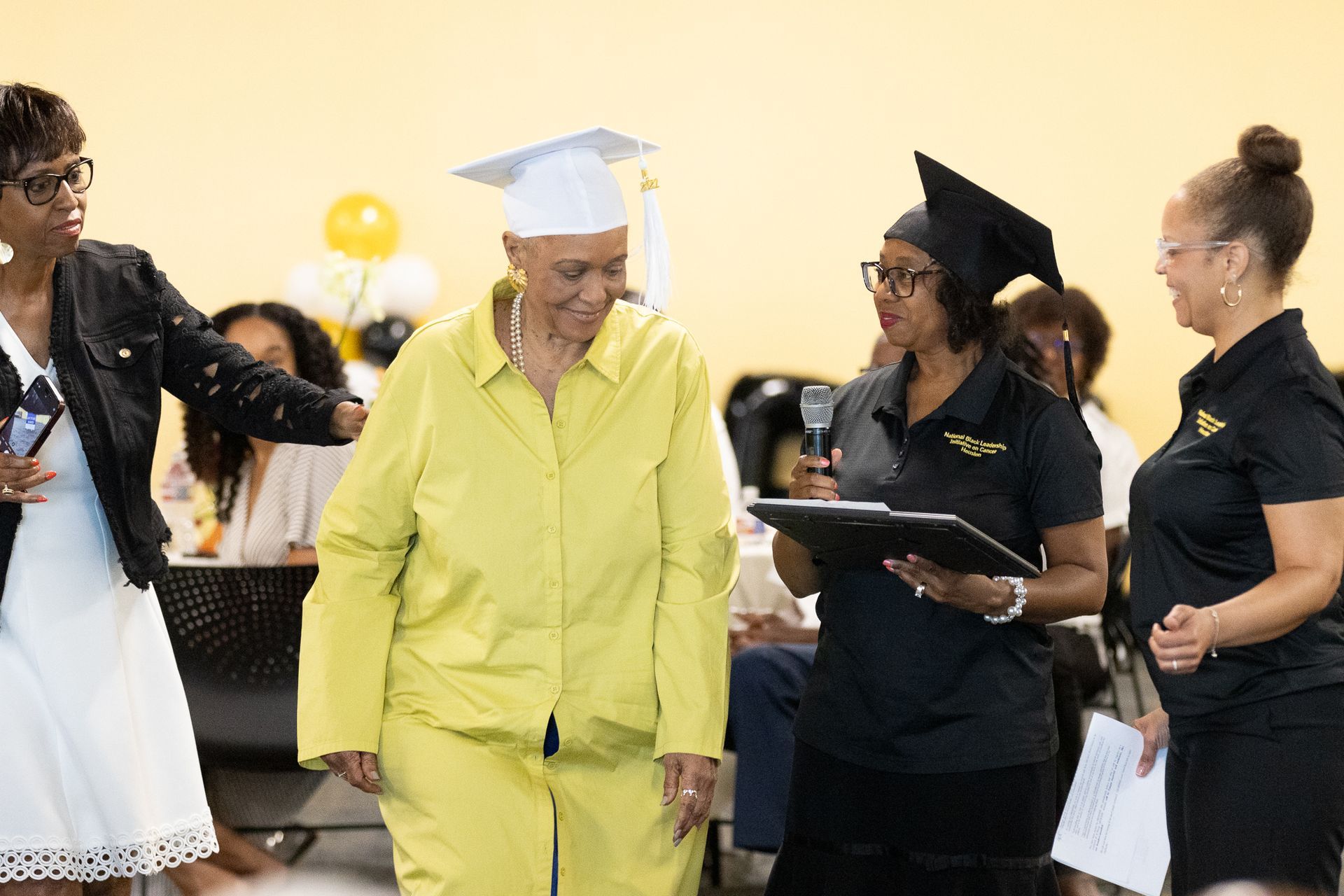 Woman in a yellow suit and graduation cap being handed a certificate on stage. People applaud.