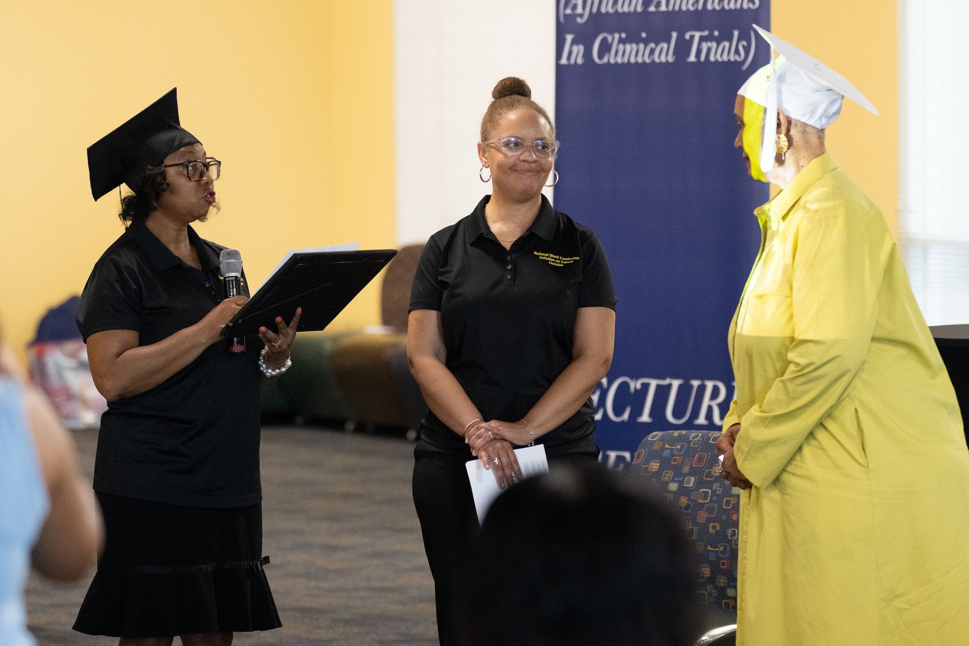 Two women and a graduate in a yellow gown at a ceremony. The speaker reads from a folder.