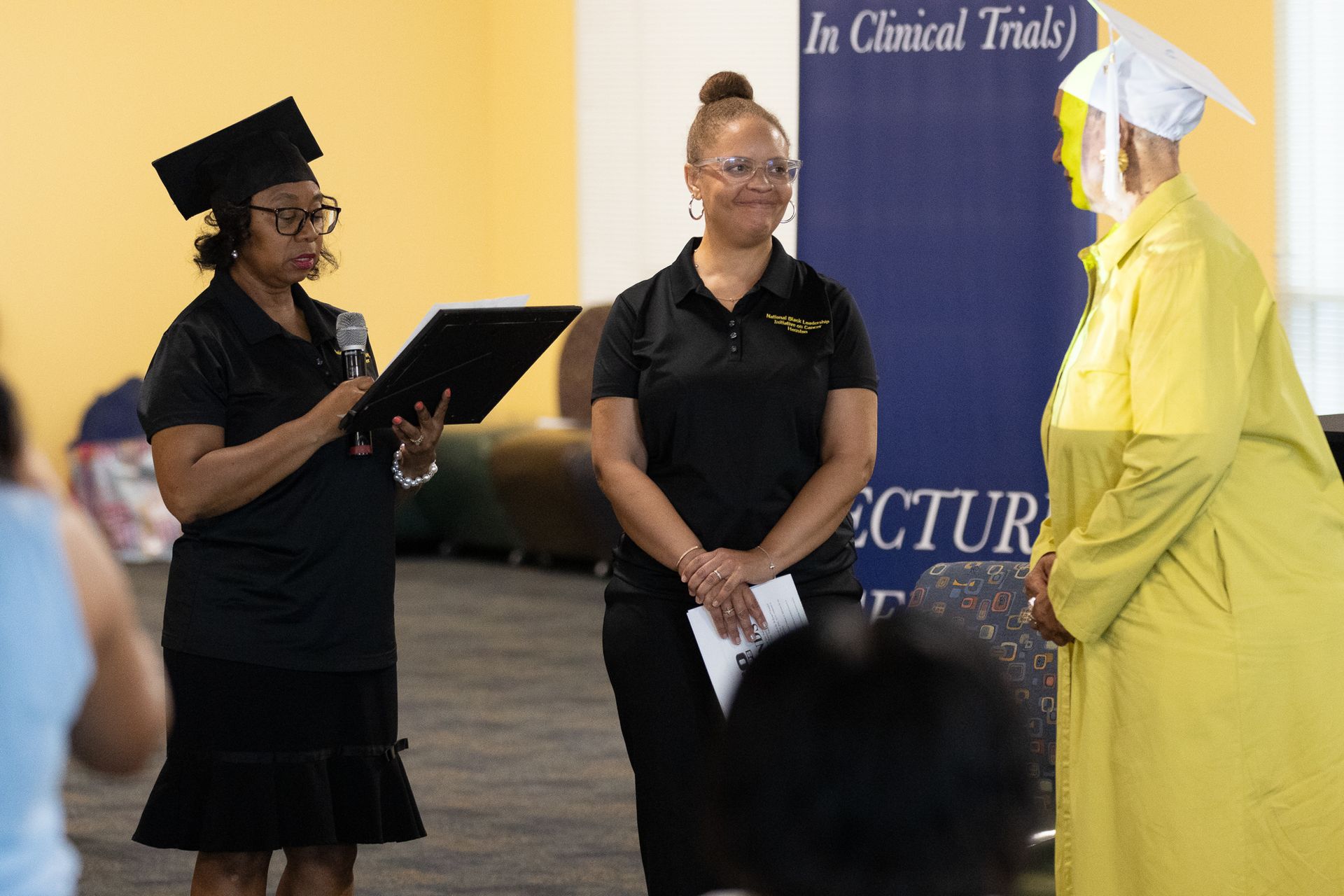 Woman in cap reads to another in yellow gown. Two others in black shirts look on.