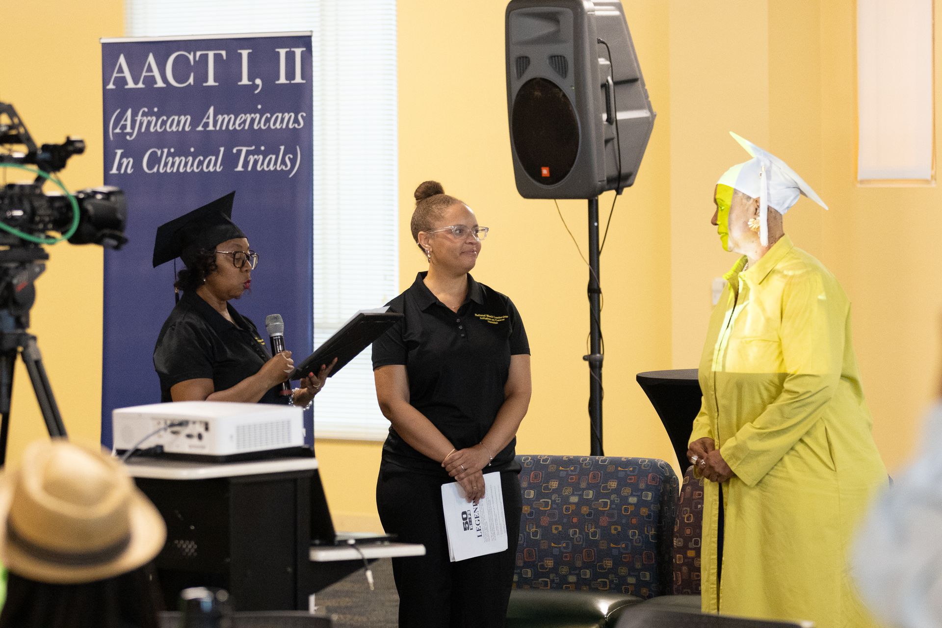A person in a yellow graduation robe receives a diploma. Ceremony with presentation screen.