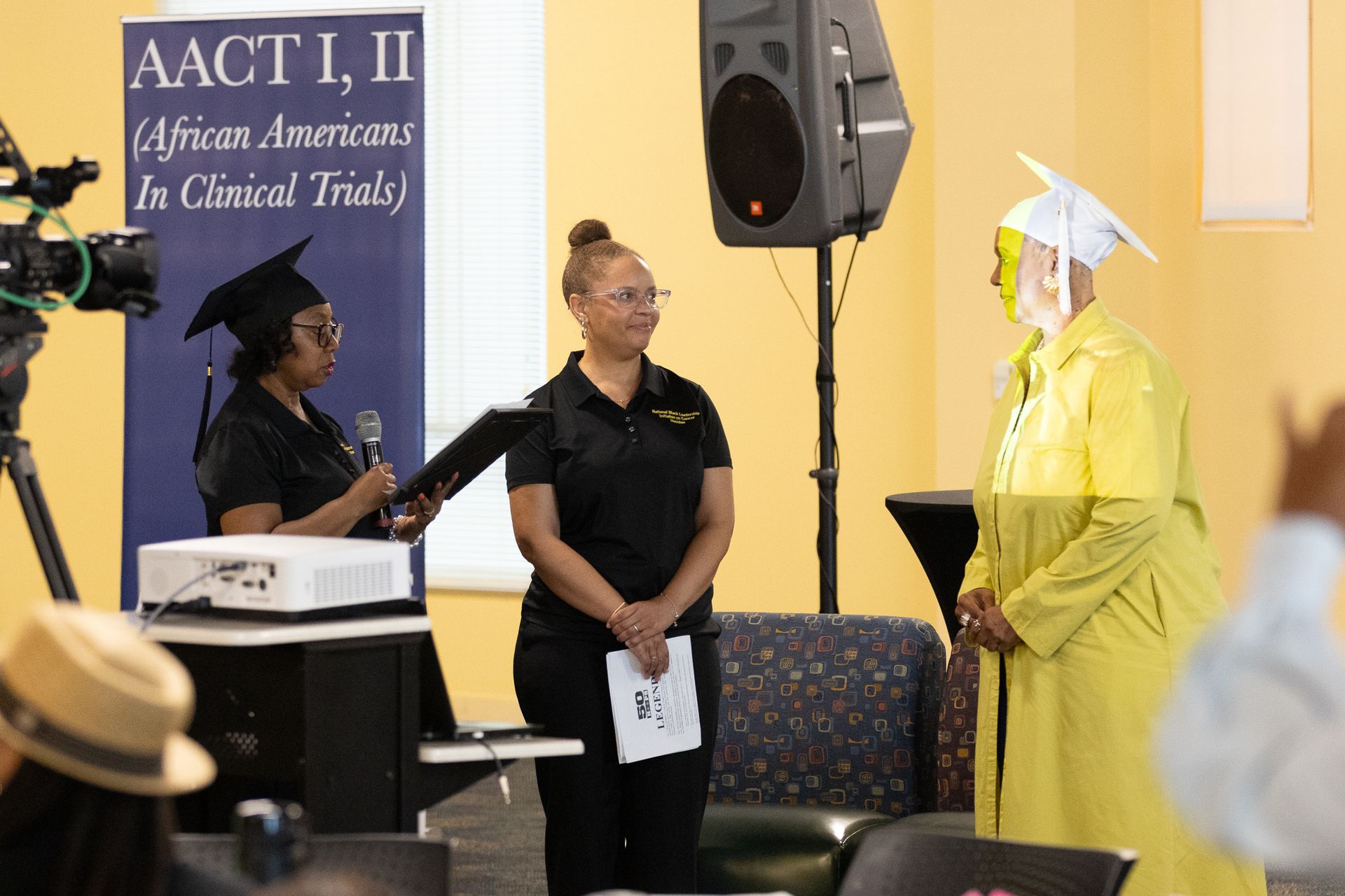 People at a presentation: two in academic caps, one in yellow robe. Banner says 