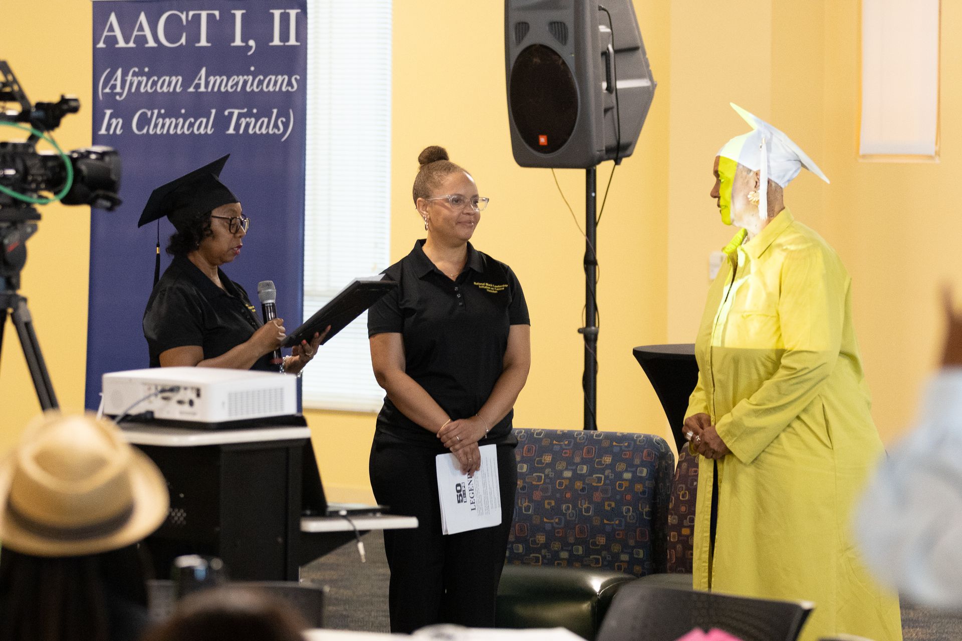 A graduation ceremony with three women. One in a cap and gown, others in black shirts. Blue banner in background.