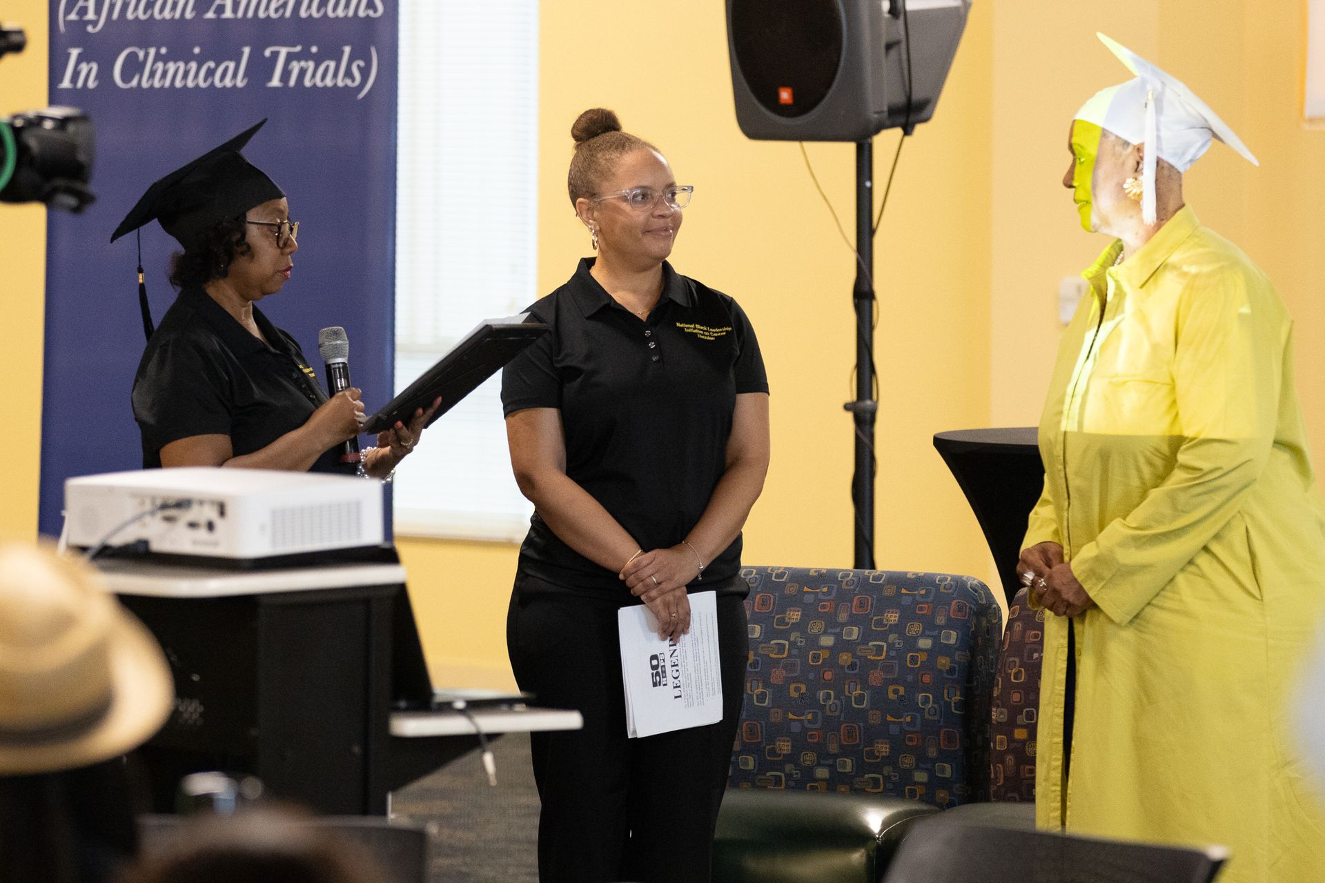 Three people in a room; one speaking, one standing holding papers, and one in graduation attire.