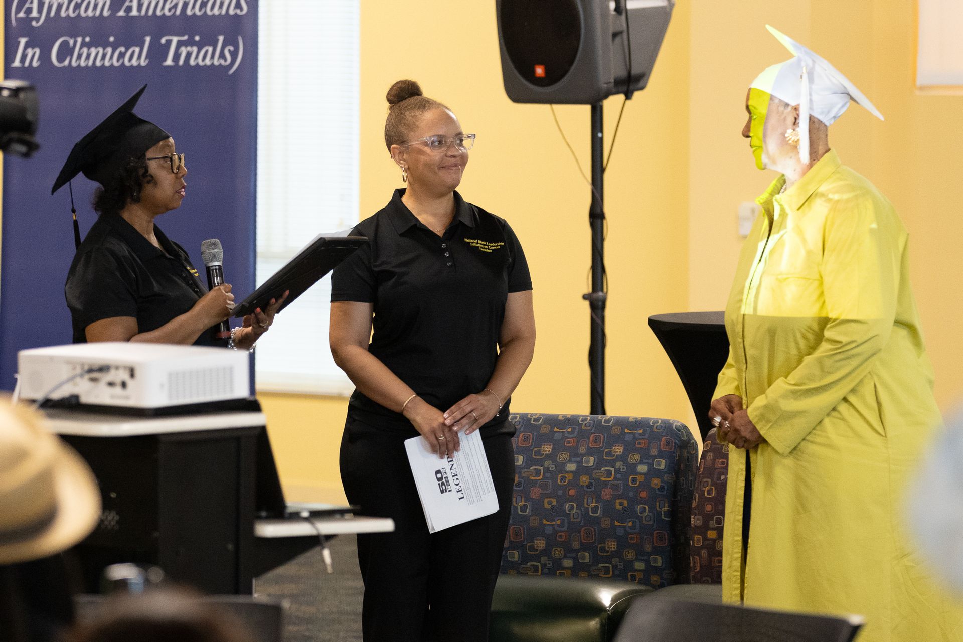 Woman in black shirt stands as another in yellow robe receives a diploma; ceremony setting.