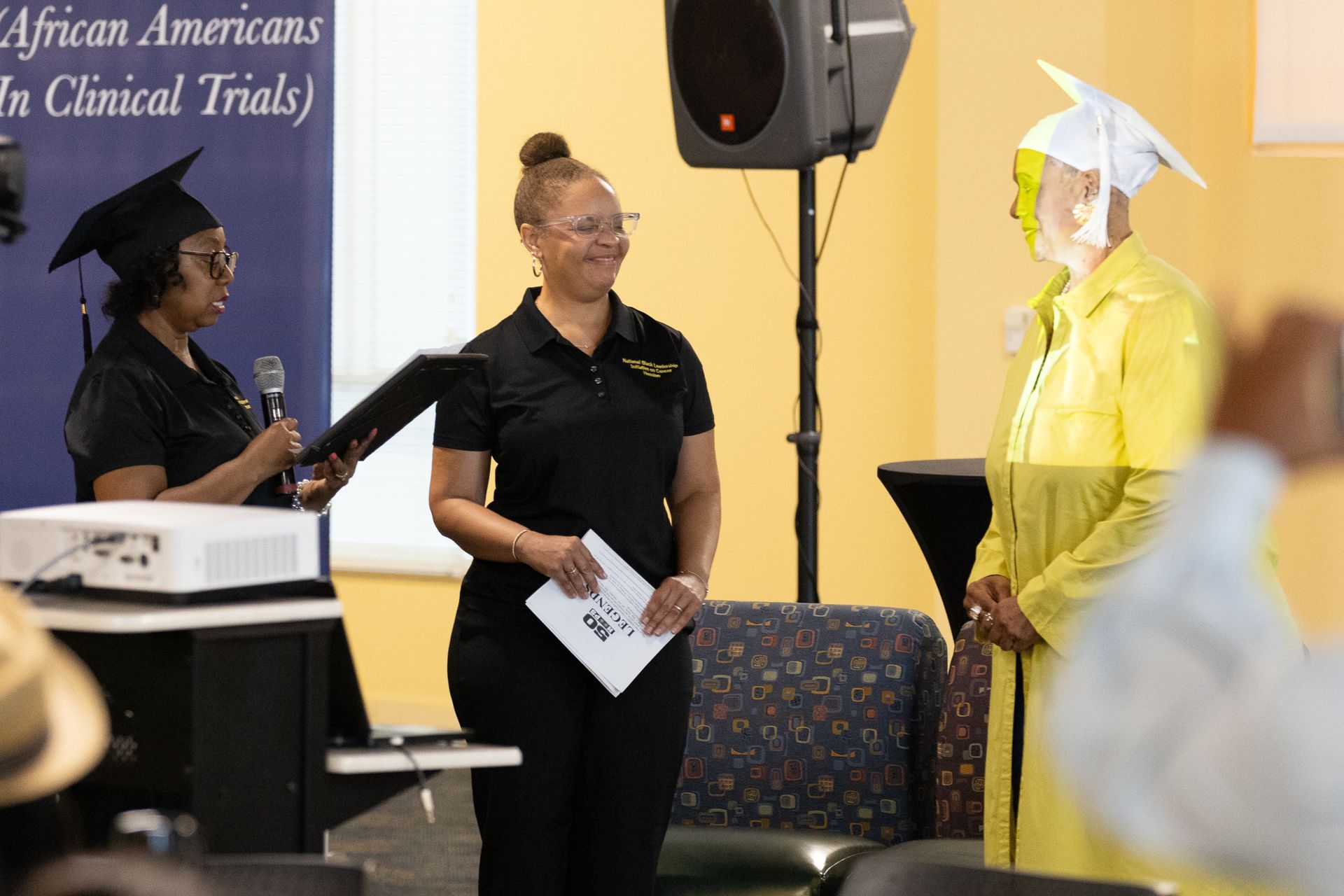 Graduation ceremony: woman in yellow gown receives certificate from two others, banner in background.