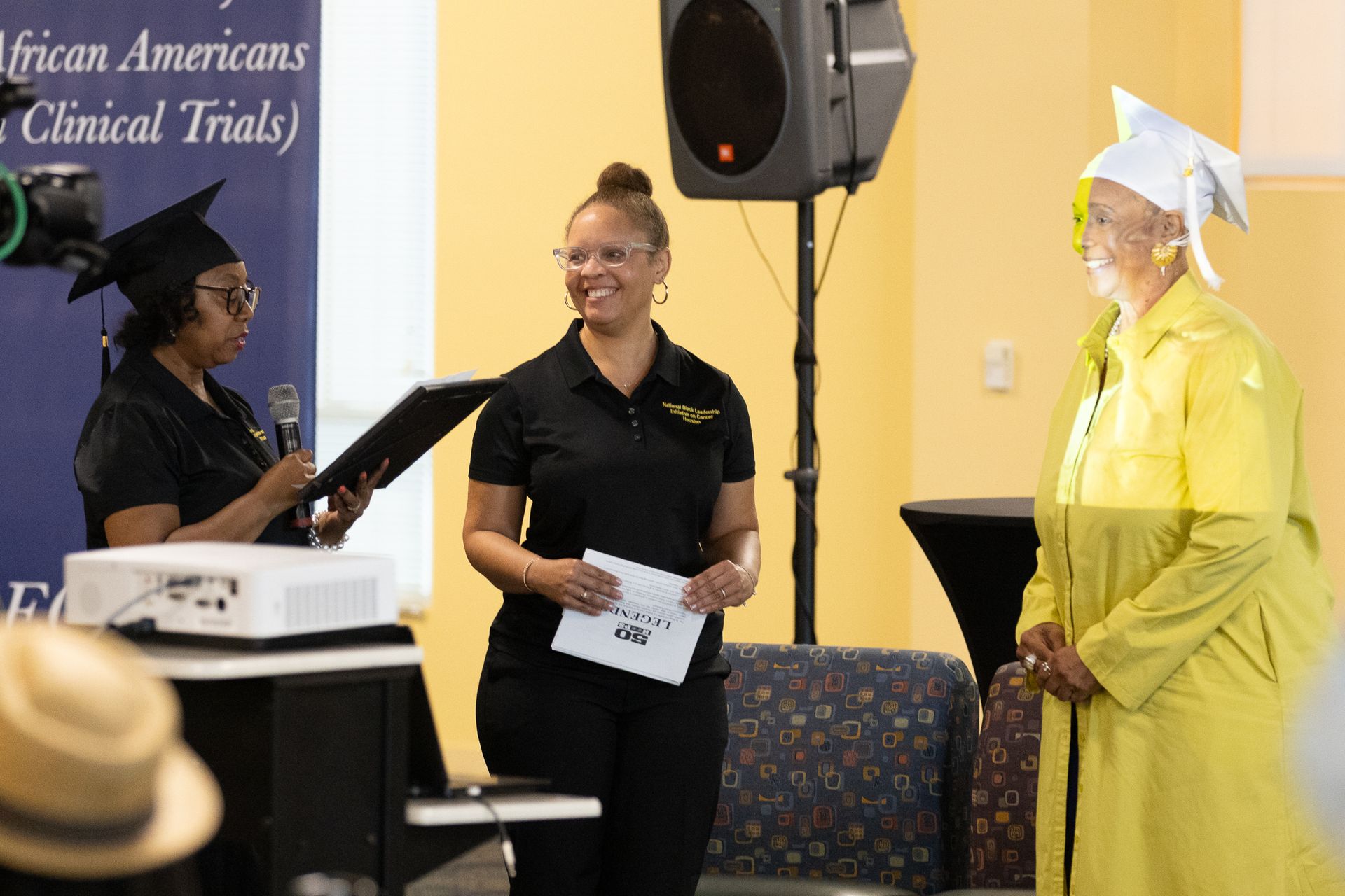 Three women at an event: one in a graduation cap reads, another holds papers, and the third stands in yellow attire.